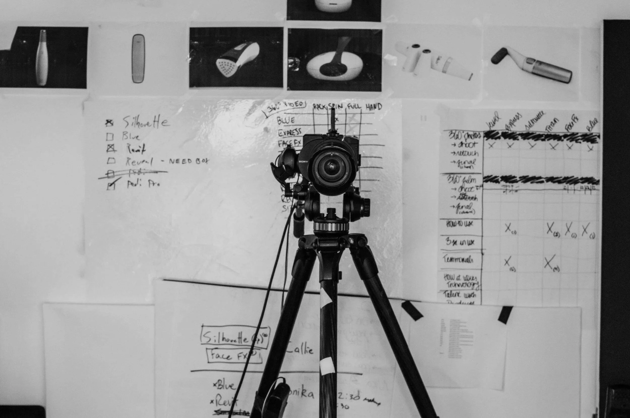 A camera on a tripod facing a whiteboard covered with handwritten notes, diagrams, and printed images of various audio equipment and production settings.