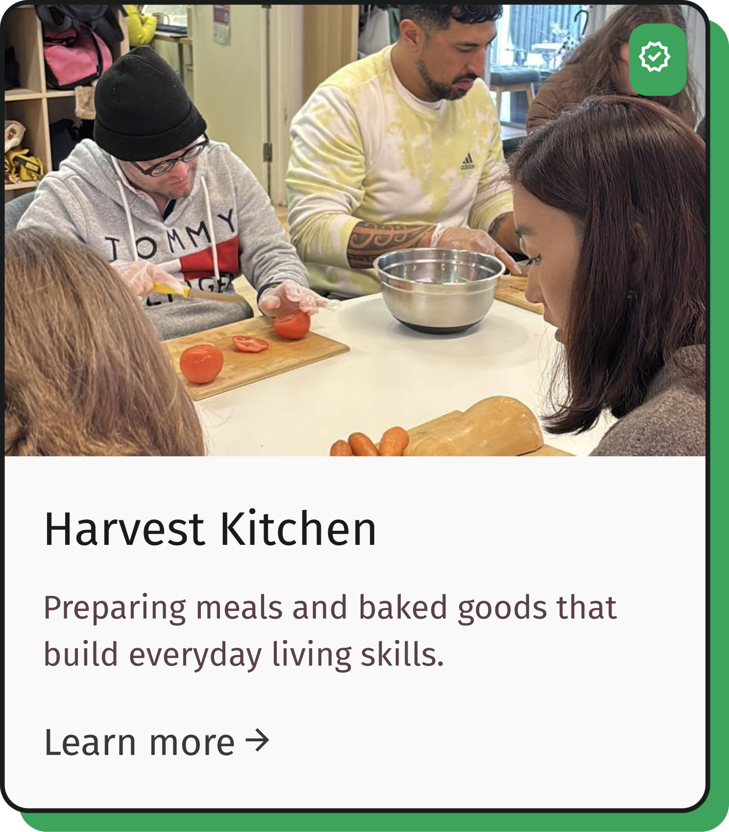 People gathered around a table preparing food in a kitchen, with tomatoes and carrots on the table.
