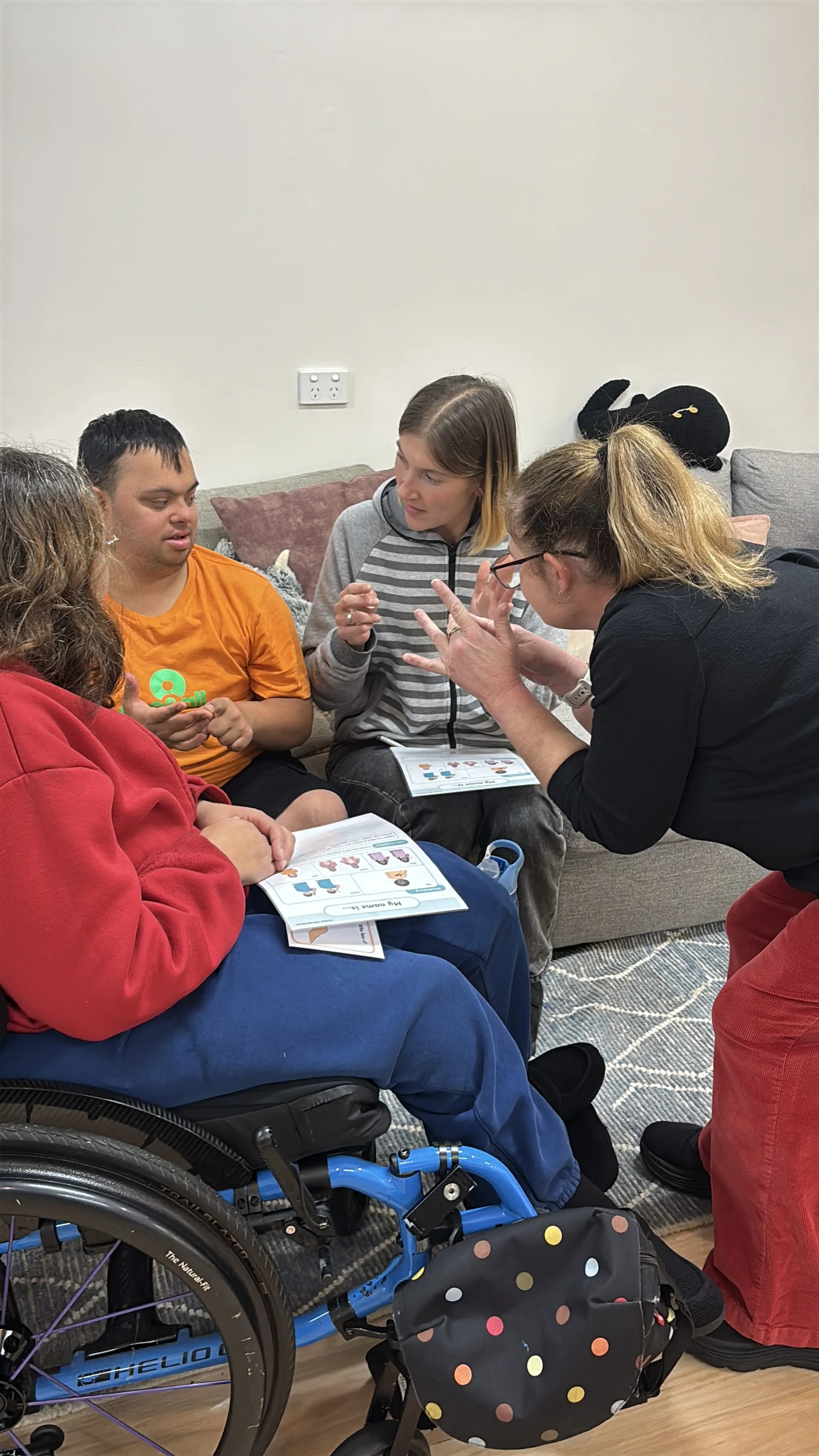 A group of four diverse people, including a person in a wheelchair, are sitting on a couch and engaging in a discussion or activity. They are holding booklets or papers with colorful illustrations, and one woman appears to be explaining something to the others. The setting is a cozy indoor space with a plain wall and a few decorative pillows.