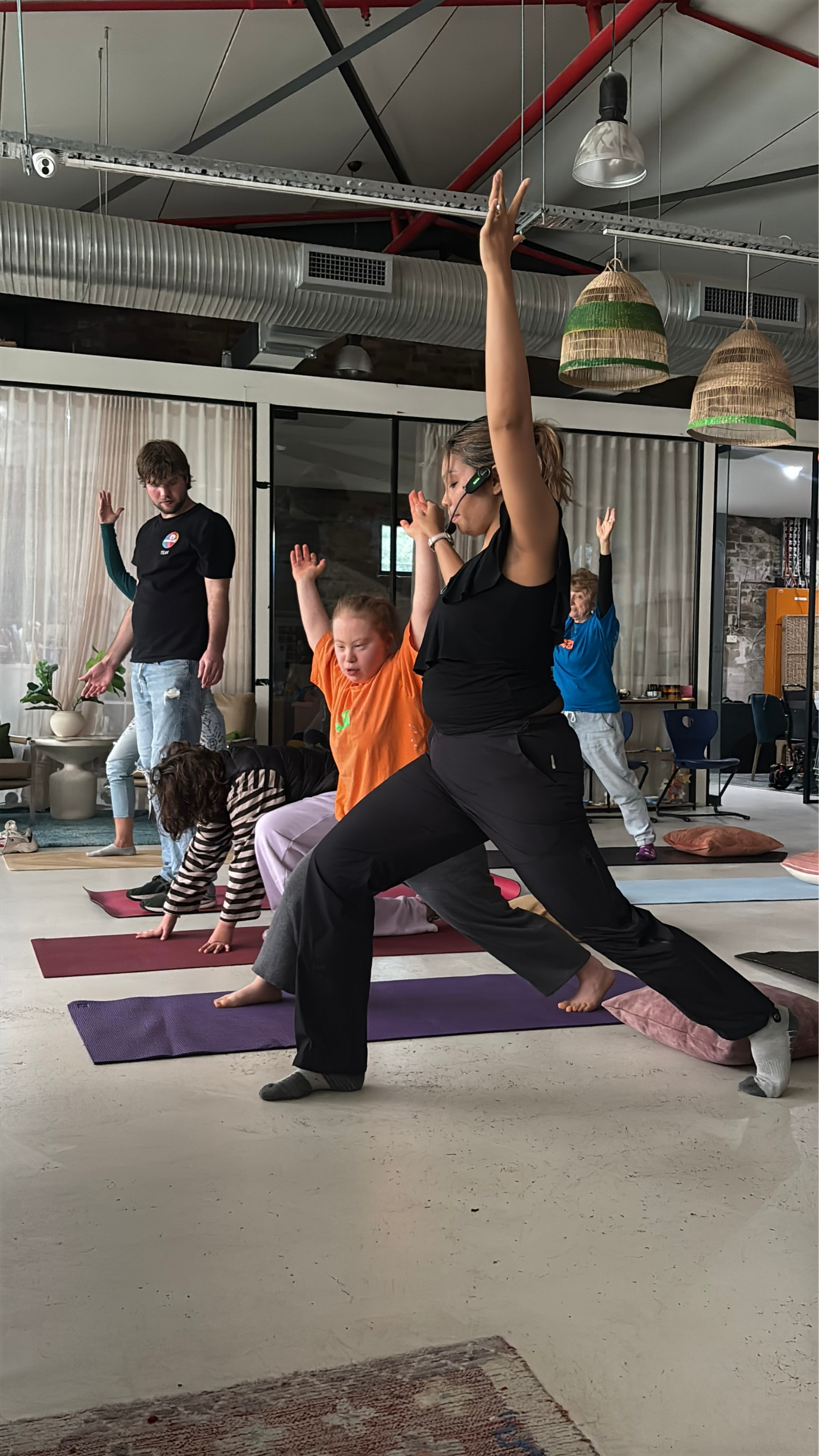Yoga class with children and instructor in a studio, instructor leading in warrior pose, children following, some in plank or other poses, colorful mats on floor, industrial-style ceiling with ducts and hanging lamps.