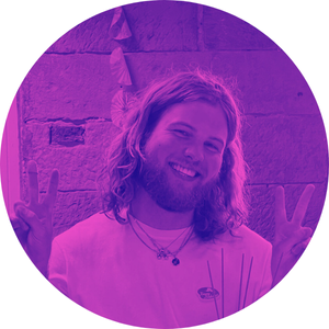 A person with long hair and a beard smiling and making peace signs with both hands, standing in front of a brick wall, purple filter applied.