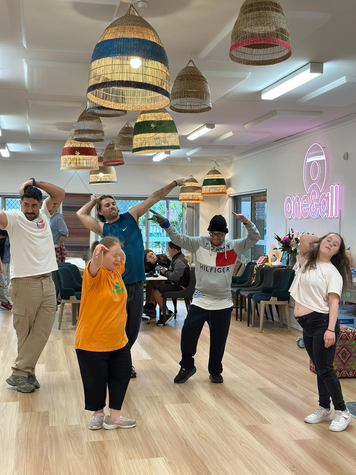 A group of five people dancing and posing inside a room with hanging wicker lamps, a neon sign that says 'one call,' and other people sitting at tables in the background.