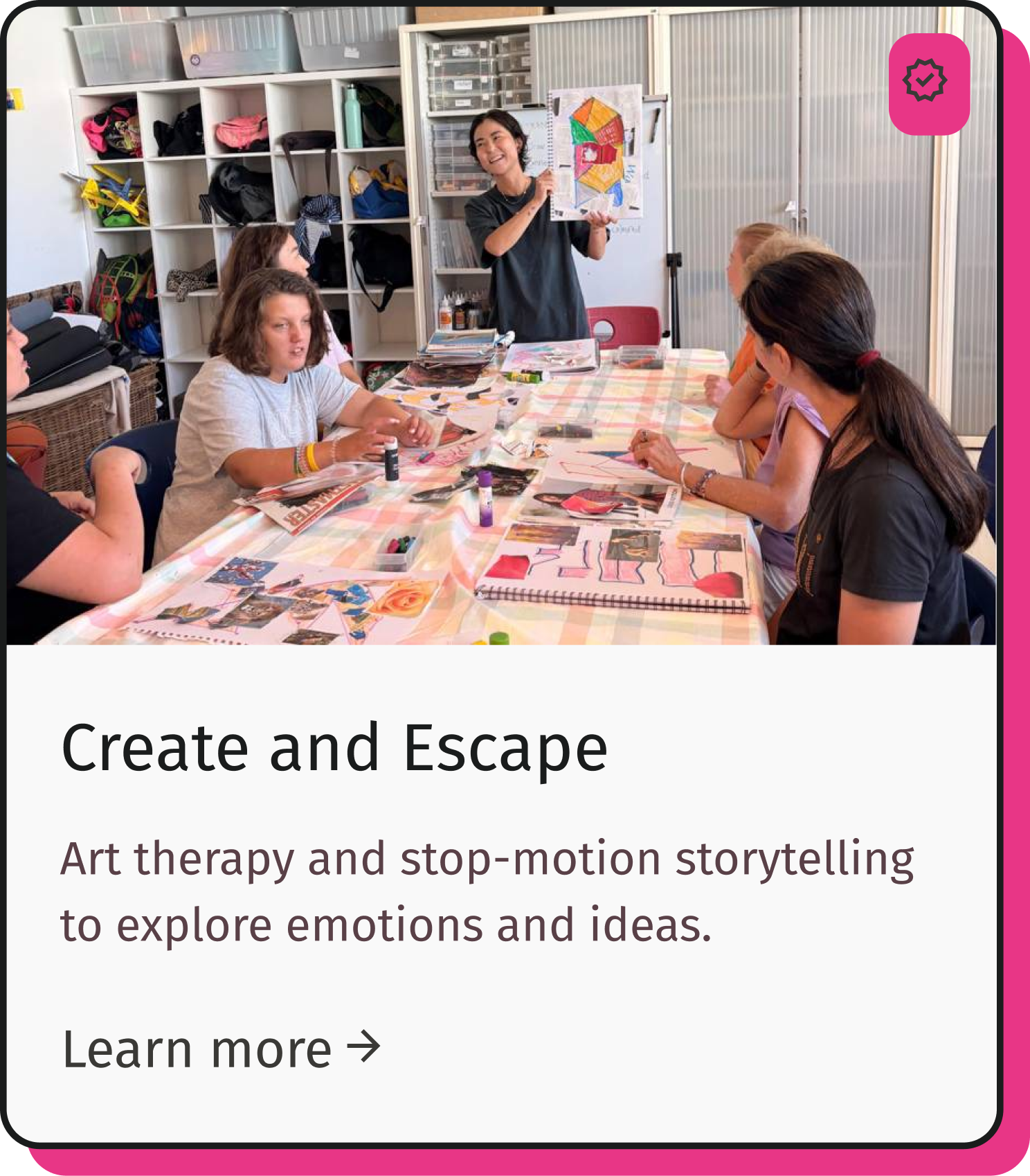 A woman stands in front of a whiteboard displaying art, showing a colorful drawing, as a group of children sit at a table covered with magazines, papers, and art supplies in a classroom setting.