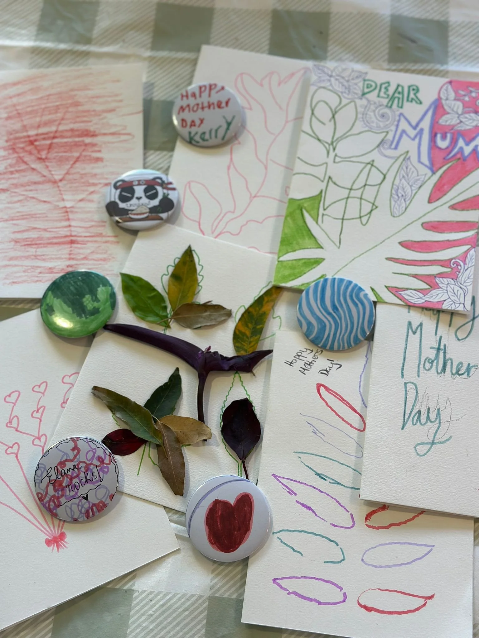 A collection of handwritten cards, drawings, and decorations celebrating Mother's Day, including illustrations of flowers, hearts, and messages such as 'Happy Mother Day Kelly' and 'Dear Mom'. There are also decorative stickers and leaves arranged among the cards.