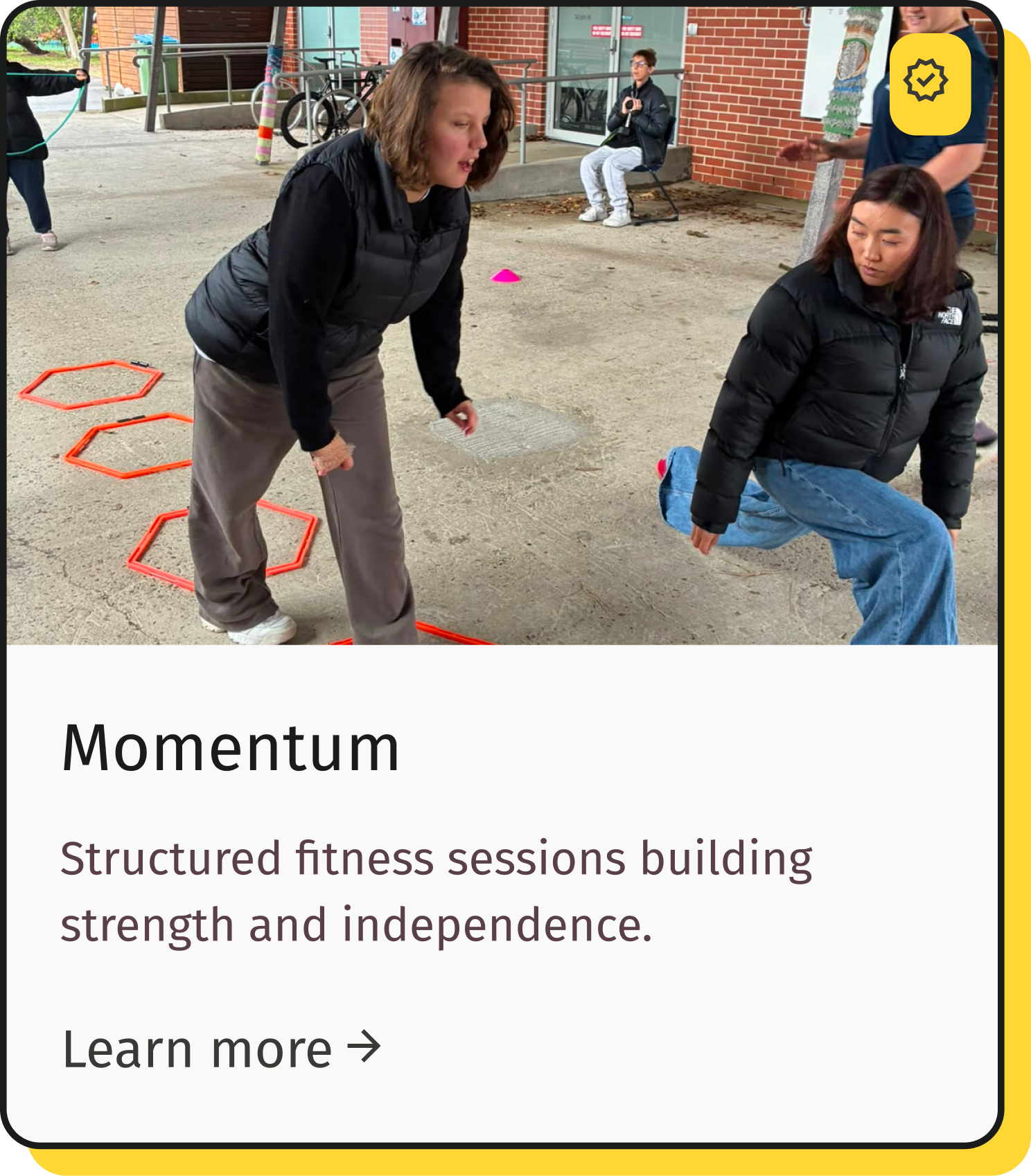 Two women participating in an outdoor fitness session, with colorful agility rings on the ground, a child in the background holding a jump rope, and a person sitting on a bench near a brick building.
