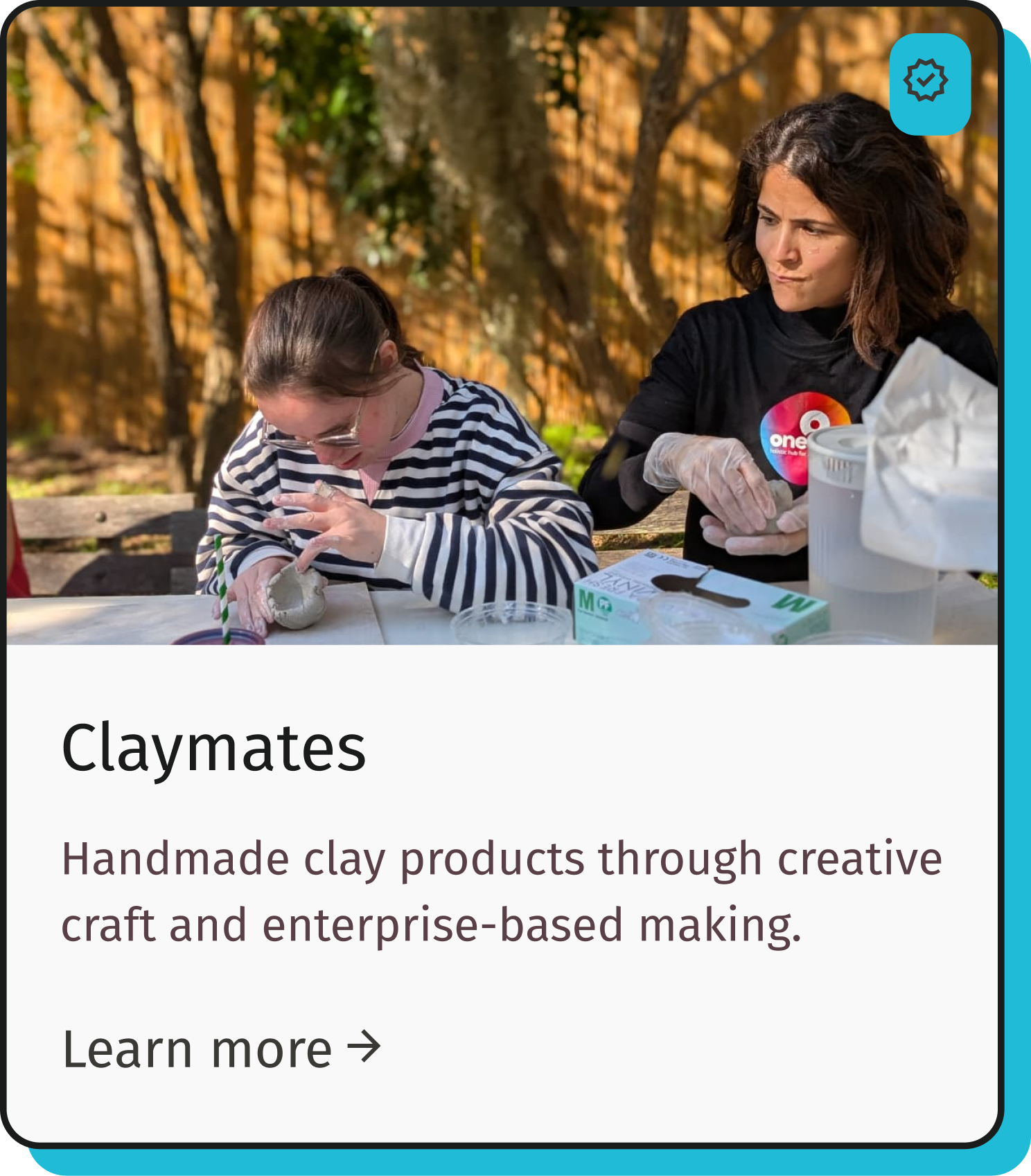 Two women working with clay outdoors, one shaping clay with her hands while the other observes, with clay and craft supplies on the table.