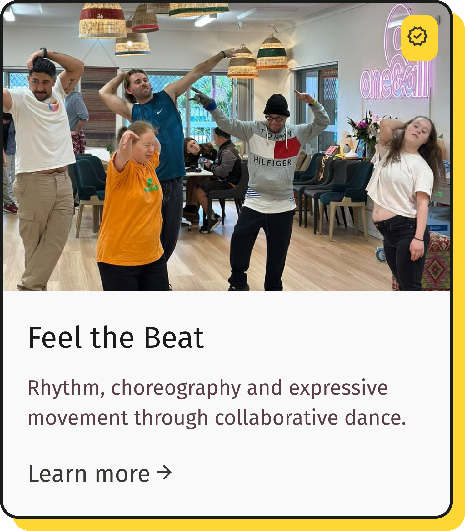 Group of people practicing dance movements in a room with colorful hanging lamps and a neon sign that says 'onecall'. They are performing rhythmic dance steps, some with arms raised and others with hands behind their heads, in a casual setting.