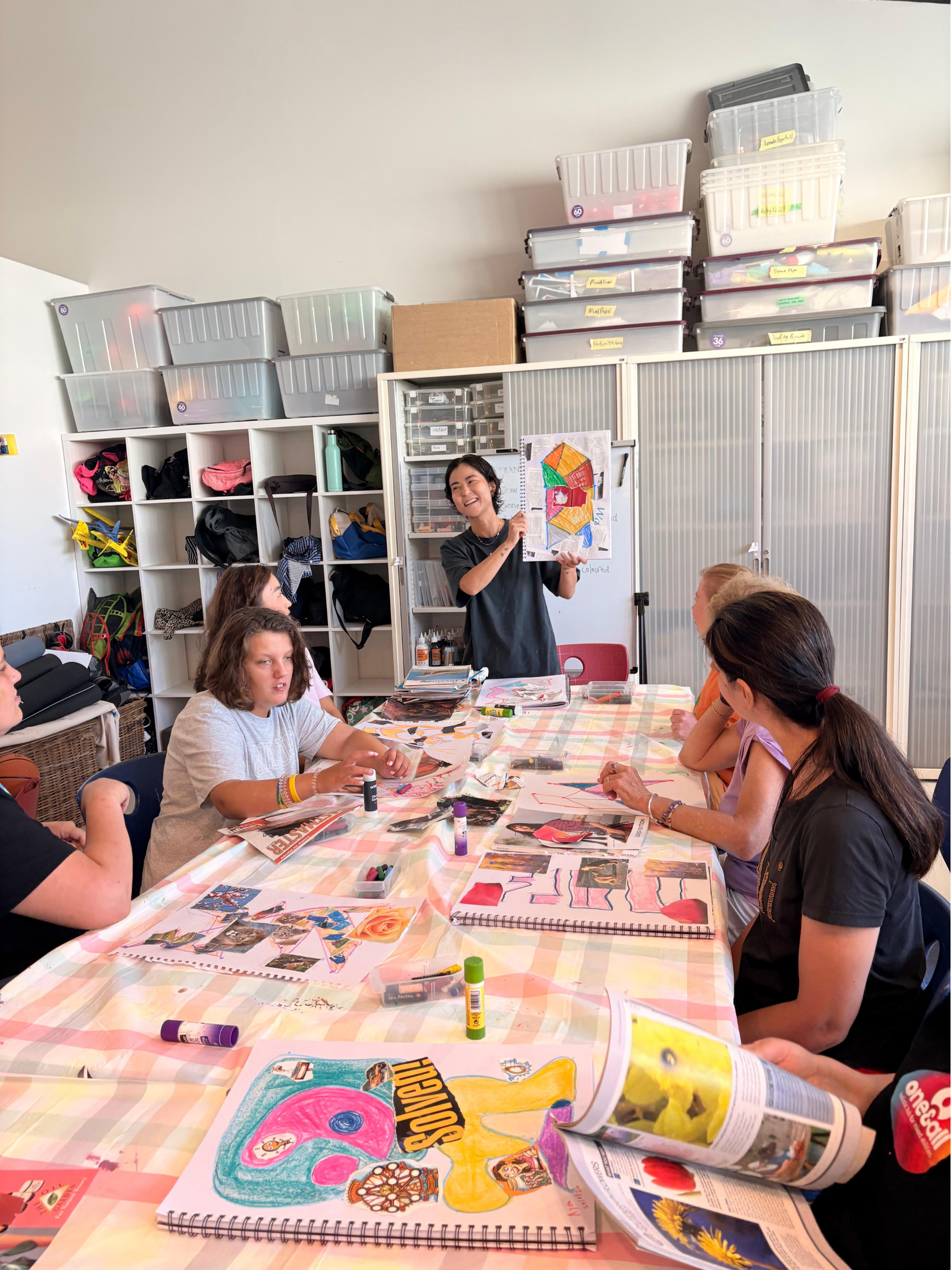 A group of people sitting around a table engaging in an arts and crafts activity, with a woman standing and holding up a colorful drawing while others work on their projects.