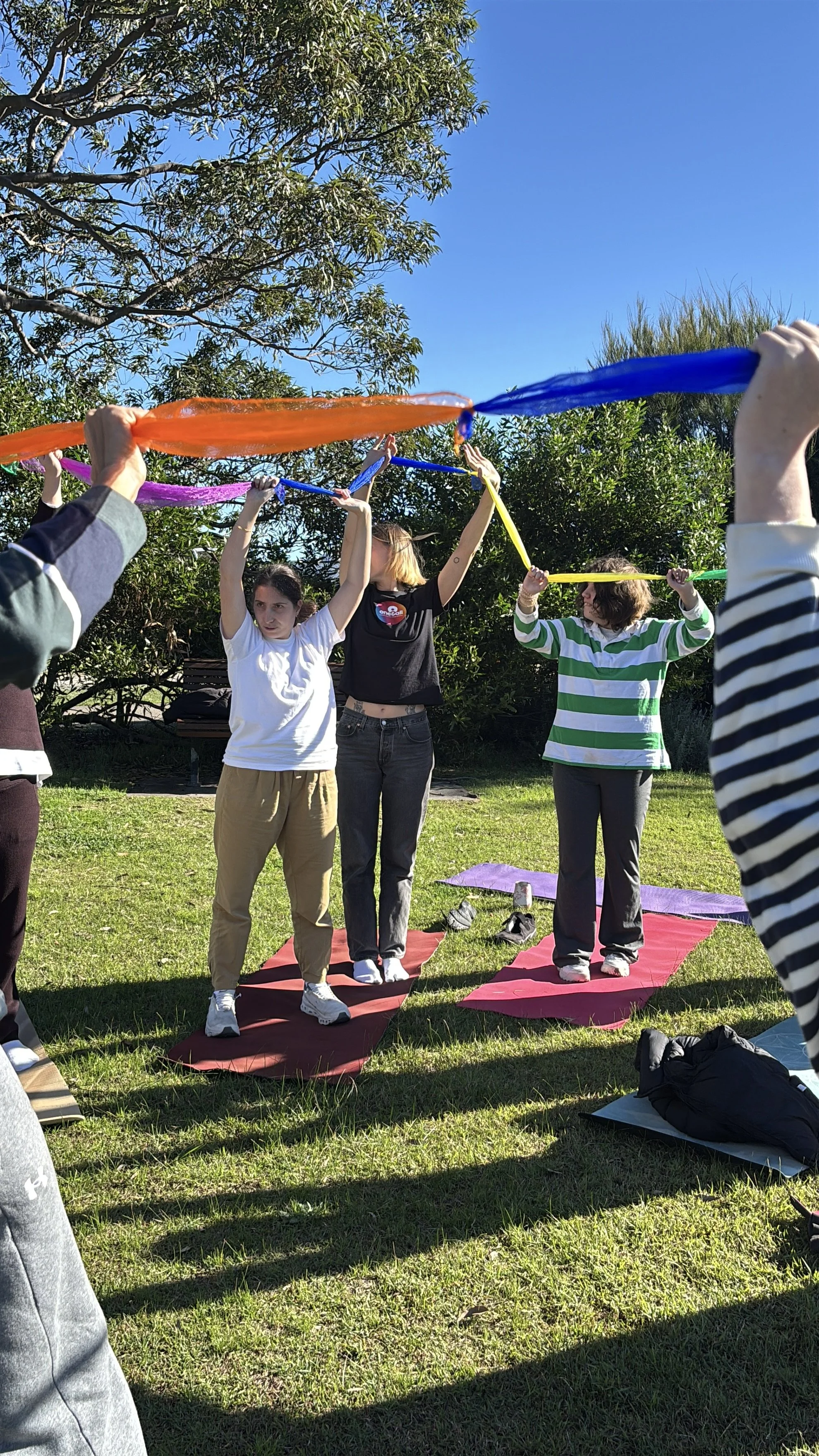 People practicing yoga outdoors on mats with colorful scarves during sunny day.