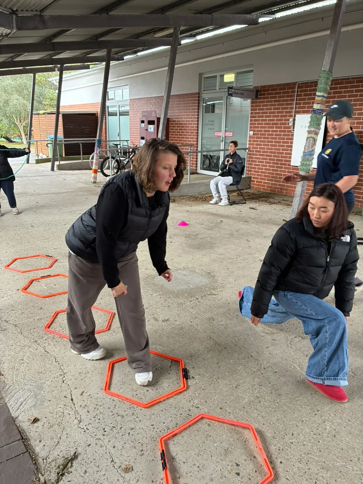 People exercising outdoors, including two women in black jackets stepping through orange agility hurdles, one woman crouching and another sitting on a bench, with a brick building in the background.