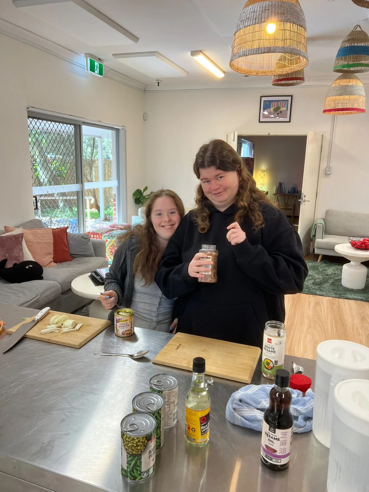 Two young women standing in a kitchen, preparing food, with cans, bottles, cutting board, and kitchen utensils on a stainless steel countertop.