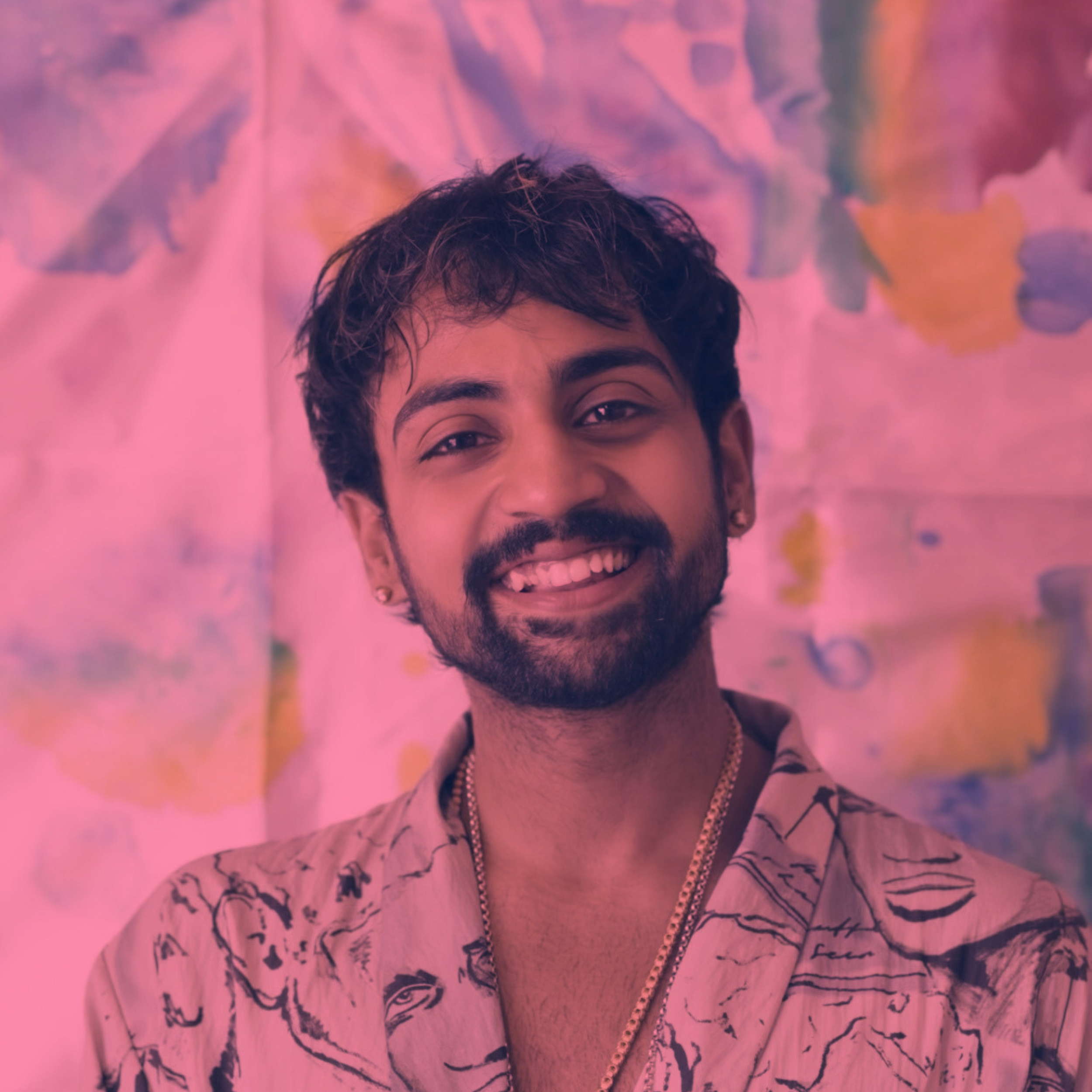 A person with short dark curly hair and a beard, smiling, wearing a patterned shirt, with a pink background featuring butterflies.