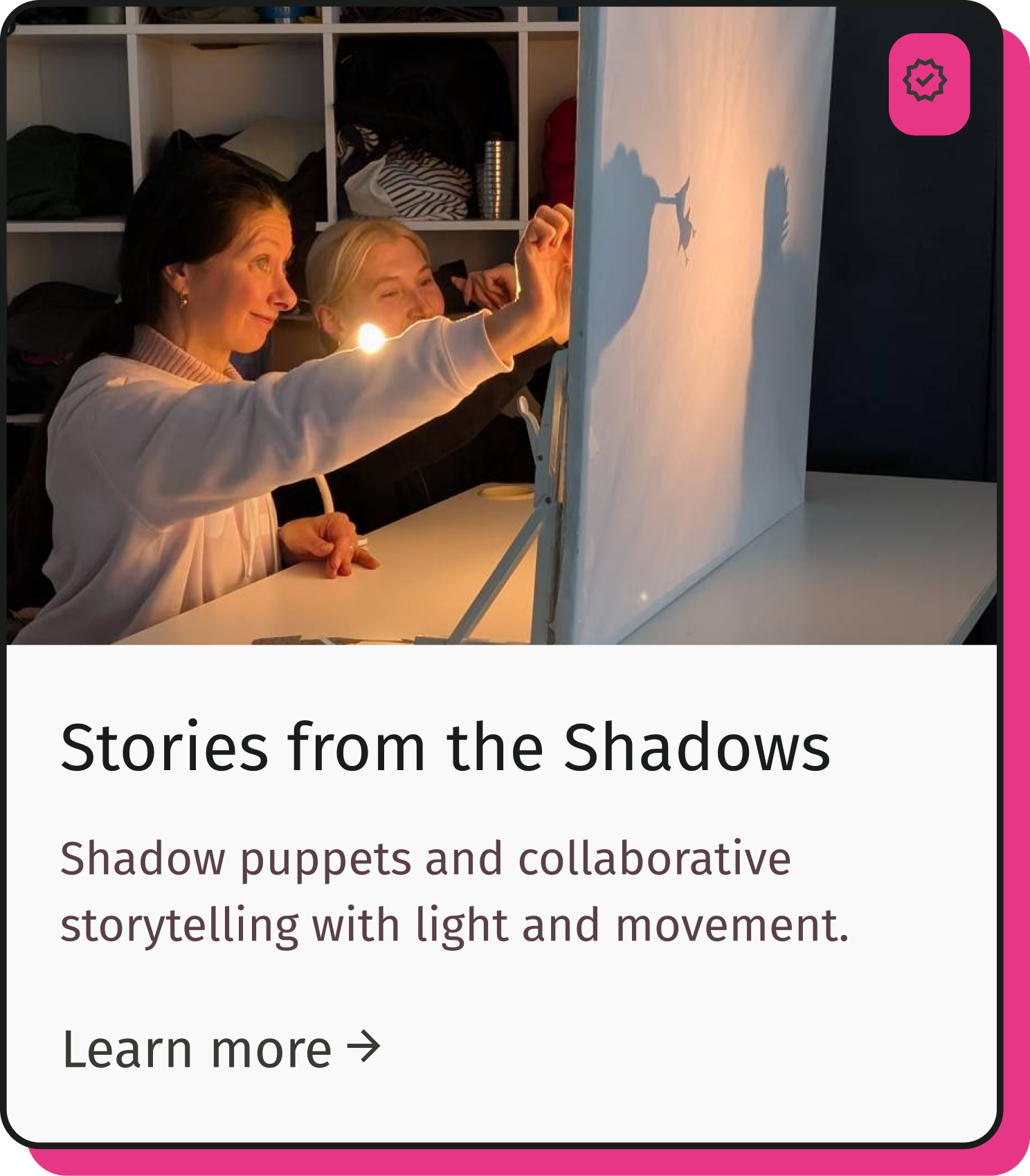 Two women sit at a table, with shadow puppets casting shadows on a screen in front of them. They are smiling and engaging in shadow puppet storytelling, with a bookshelf in the background. The scene is illuminated by warm light.