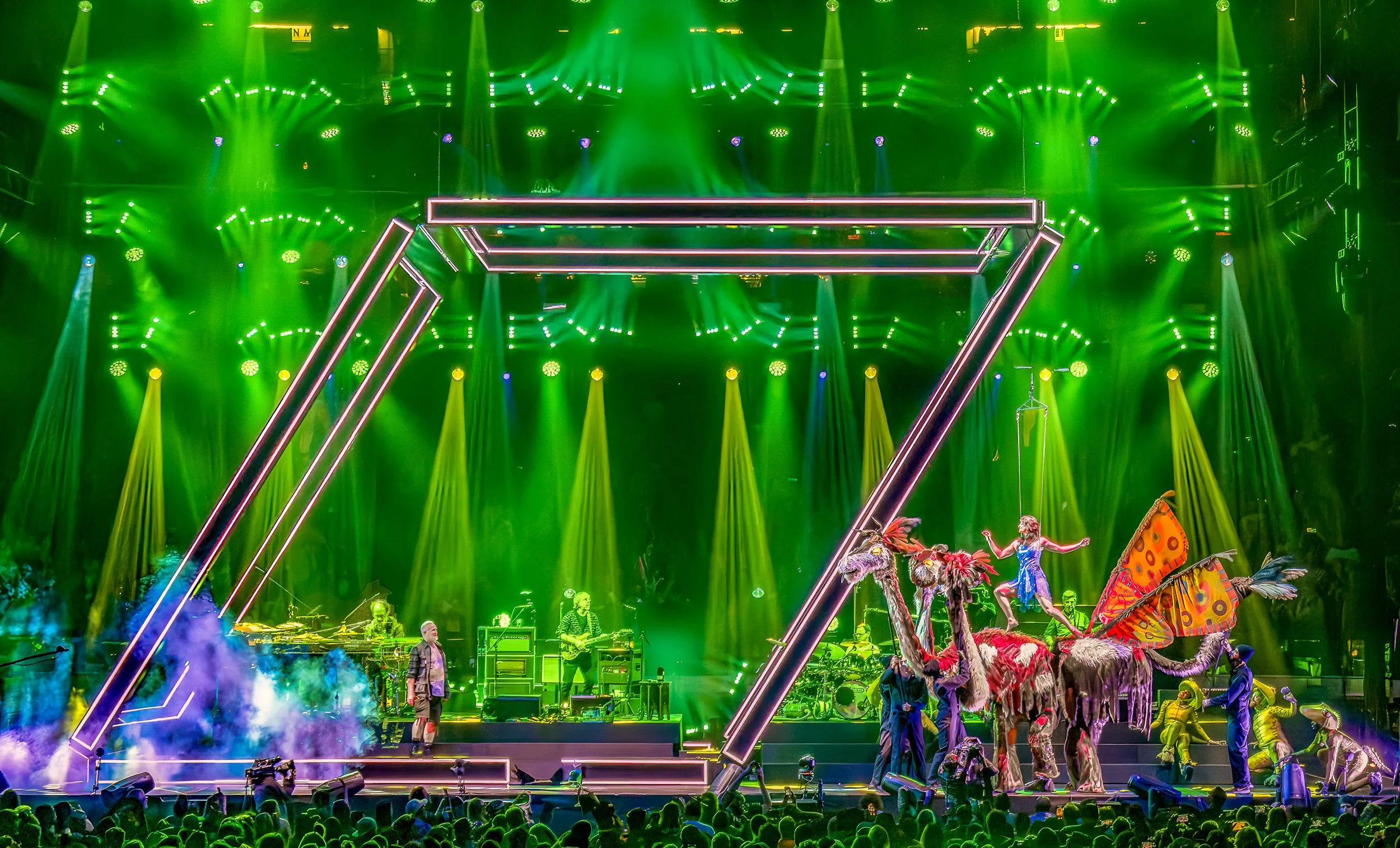 Simple Mischief Studio performing onstage with the band Phish, and their Multi-Beast puppet. Colorful stage with bright green lighting, performers in costumes, and large pink geometric structure overhead.