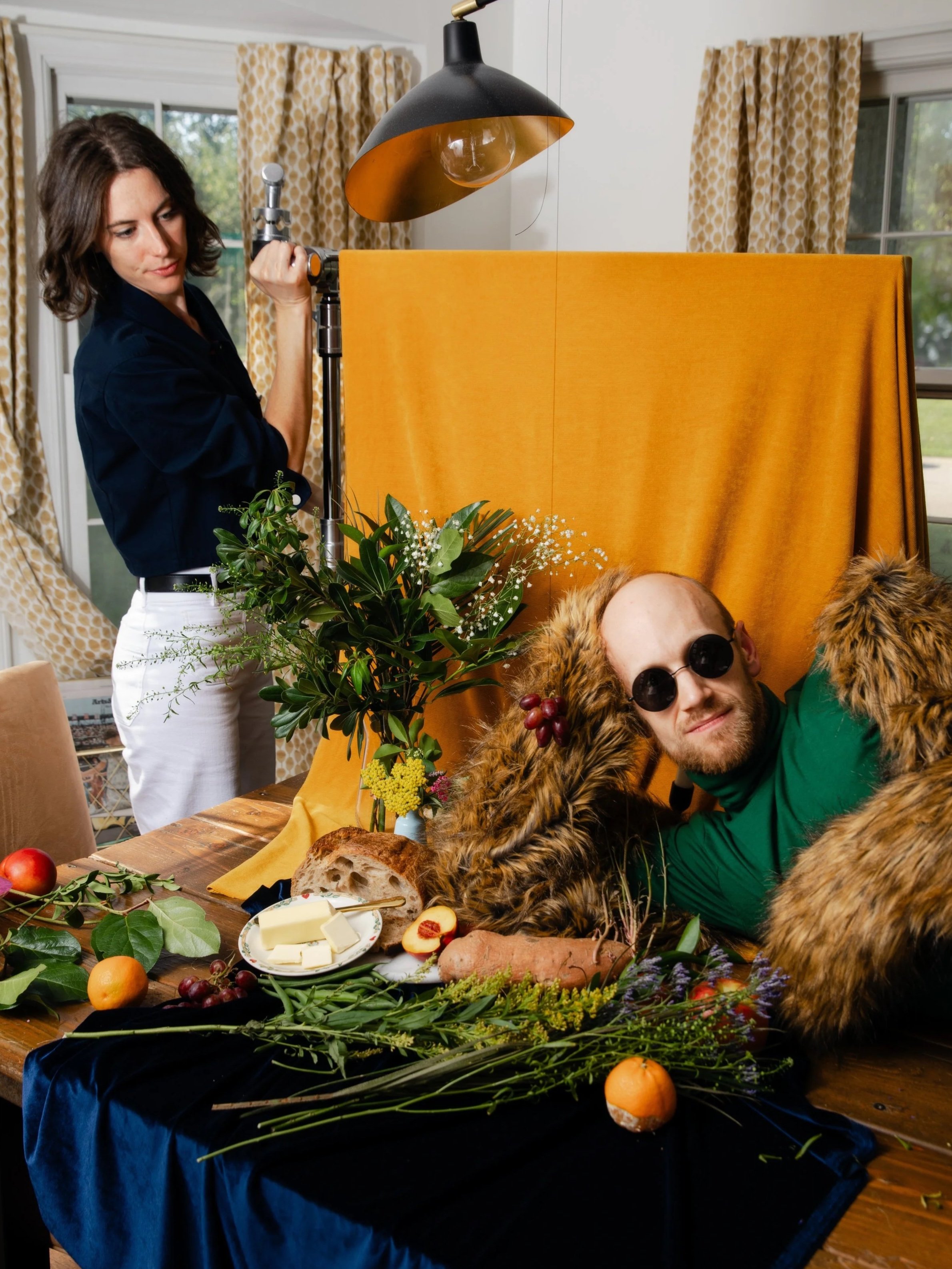 A man with sunglasses dressed as Fancy Duck, lying across a table decorated with fruits, bread, cheese, and flowers, while a woman wearing a dark top and white pants adjusts a light.
