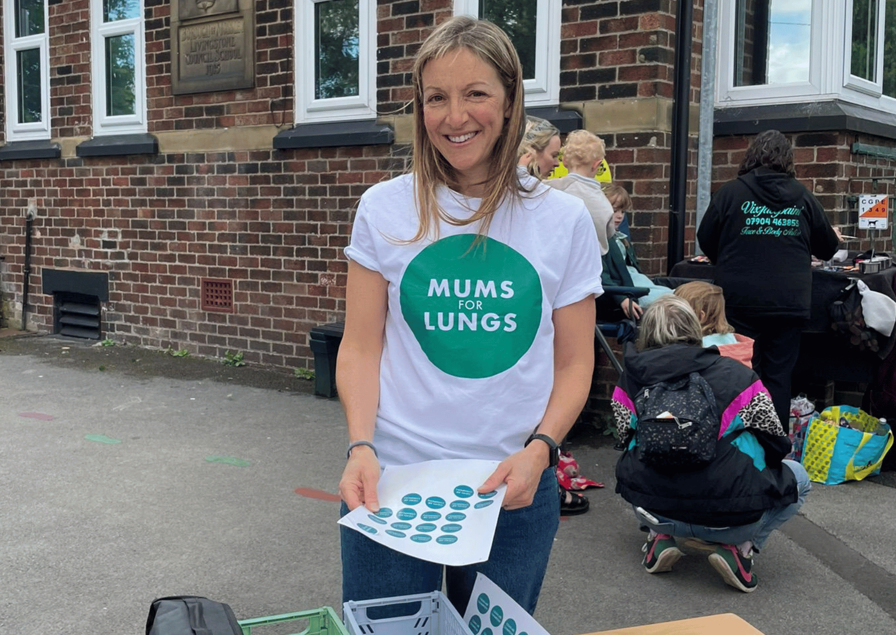 A white woman standing in a school playground in front of a table, holding some stickers.