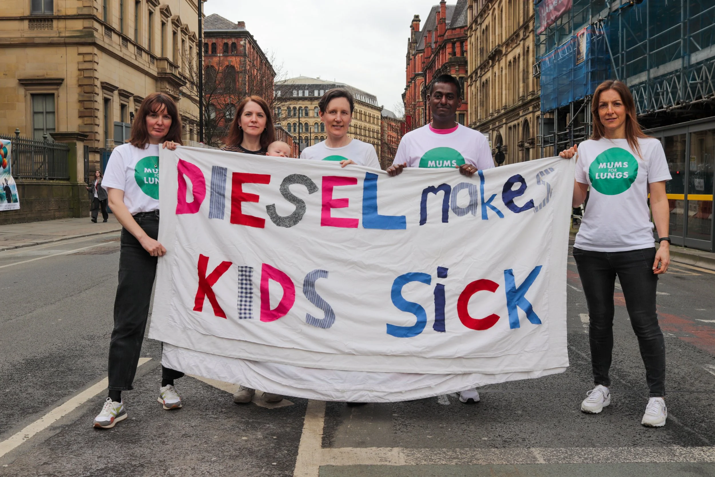 A group of people standing on a pavement, holding a banner that says "diesel makes kids sick" in colourful lettering.