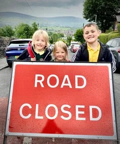 Three children standing on a road behind a big red sign that says "road closed".