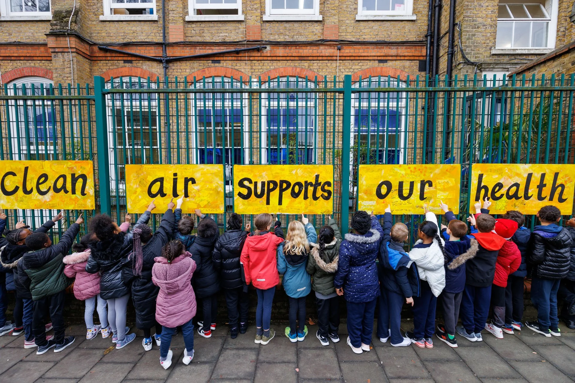 A row of children outside a school, with their backs to the camera. They are pointing at some signs they've made that say "clean air supports our health".