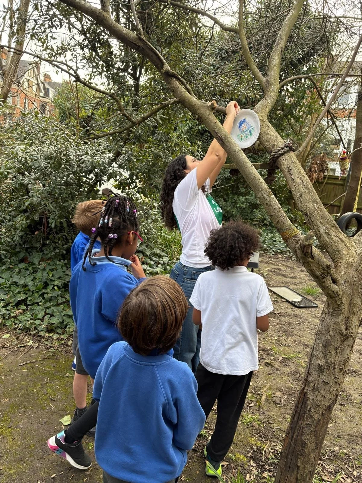 A woman in a garden, attaching a paper plate air pollution catcher to a tree. There are four children standing around her.