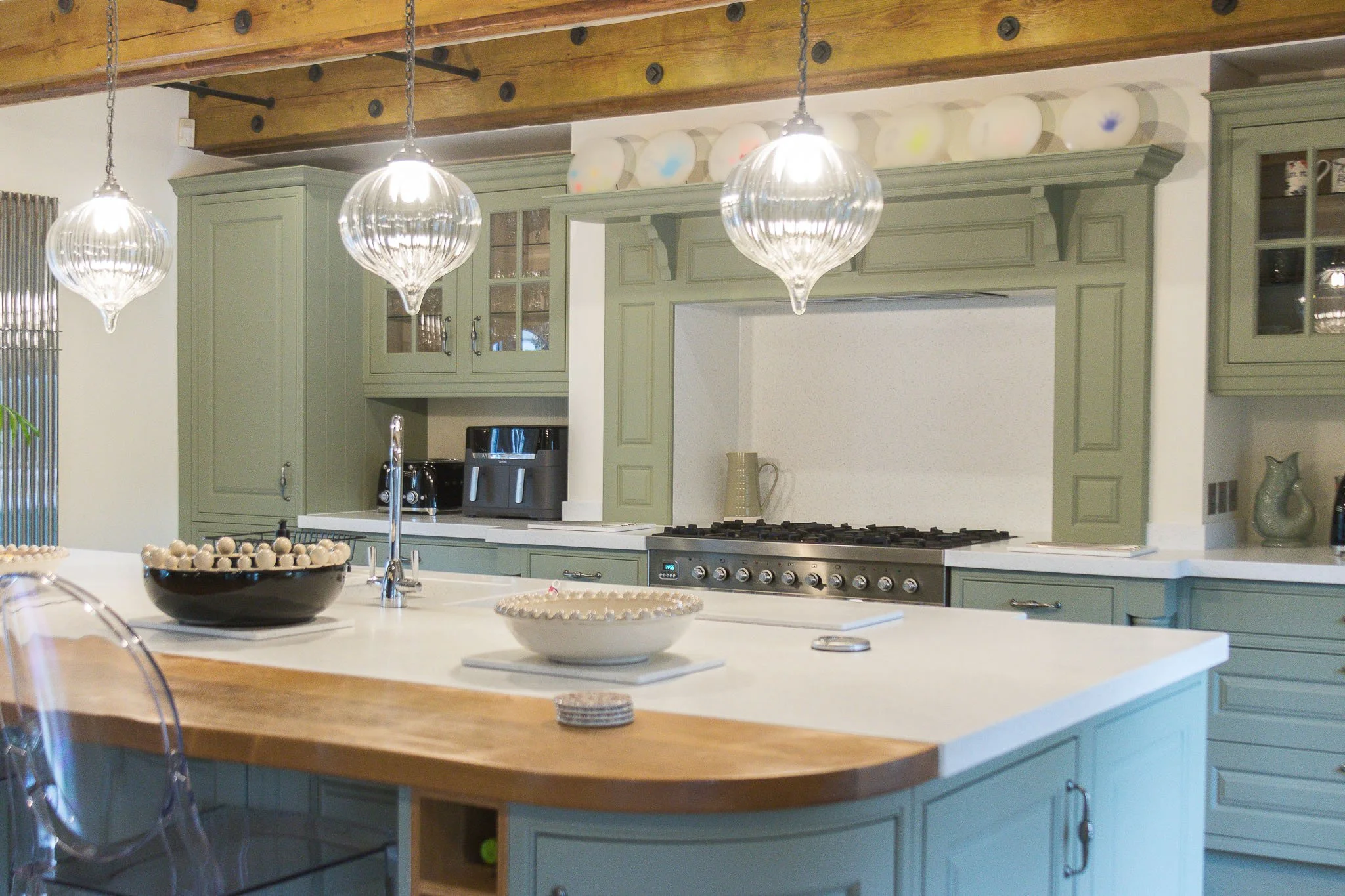 A kitchen with green cabinetry, a wooden ceiling beam, three hanging glass pendant lights, a stove, a sink, and decorative plates on a shelf above the cabinets.