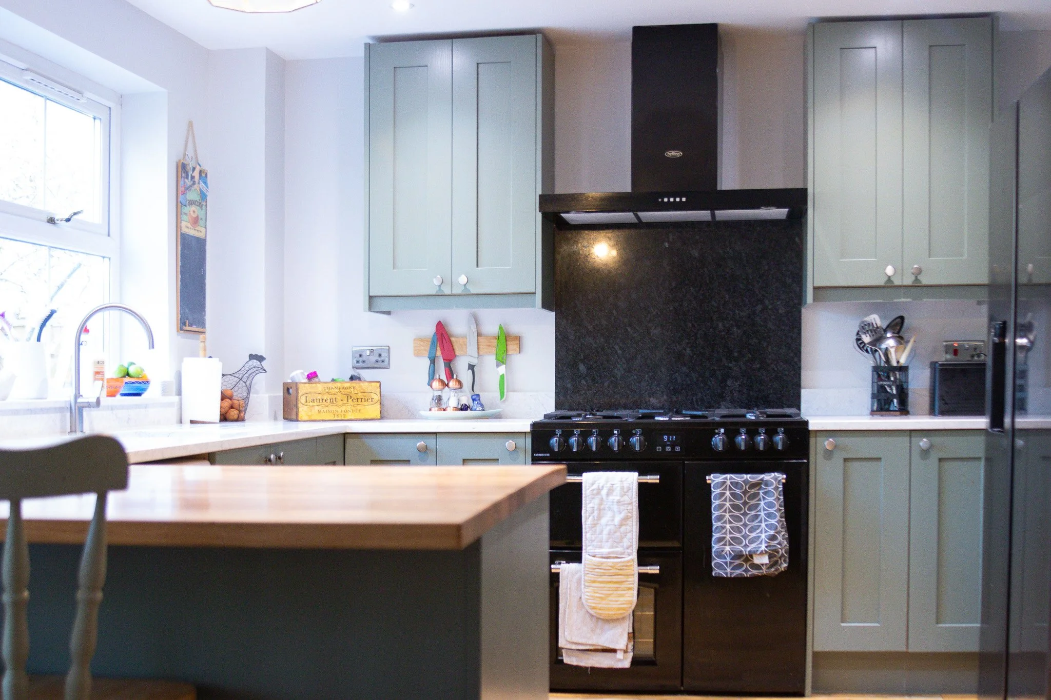 Kitchen with green cabinets, a black stove and oven, a black range hood, light-colored countertops, a window with a sink, and various kitchen utensils and decorations.