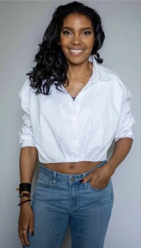 A woman with long, curly black hair, wearing a white crop top shirt and blue jeans, smiling at the camera.