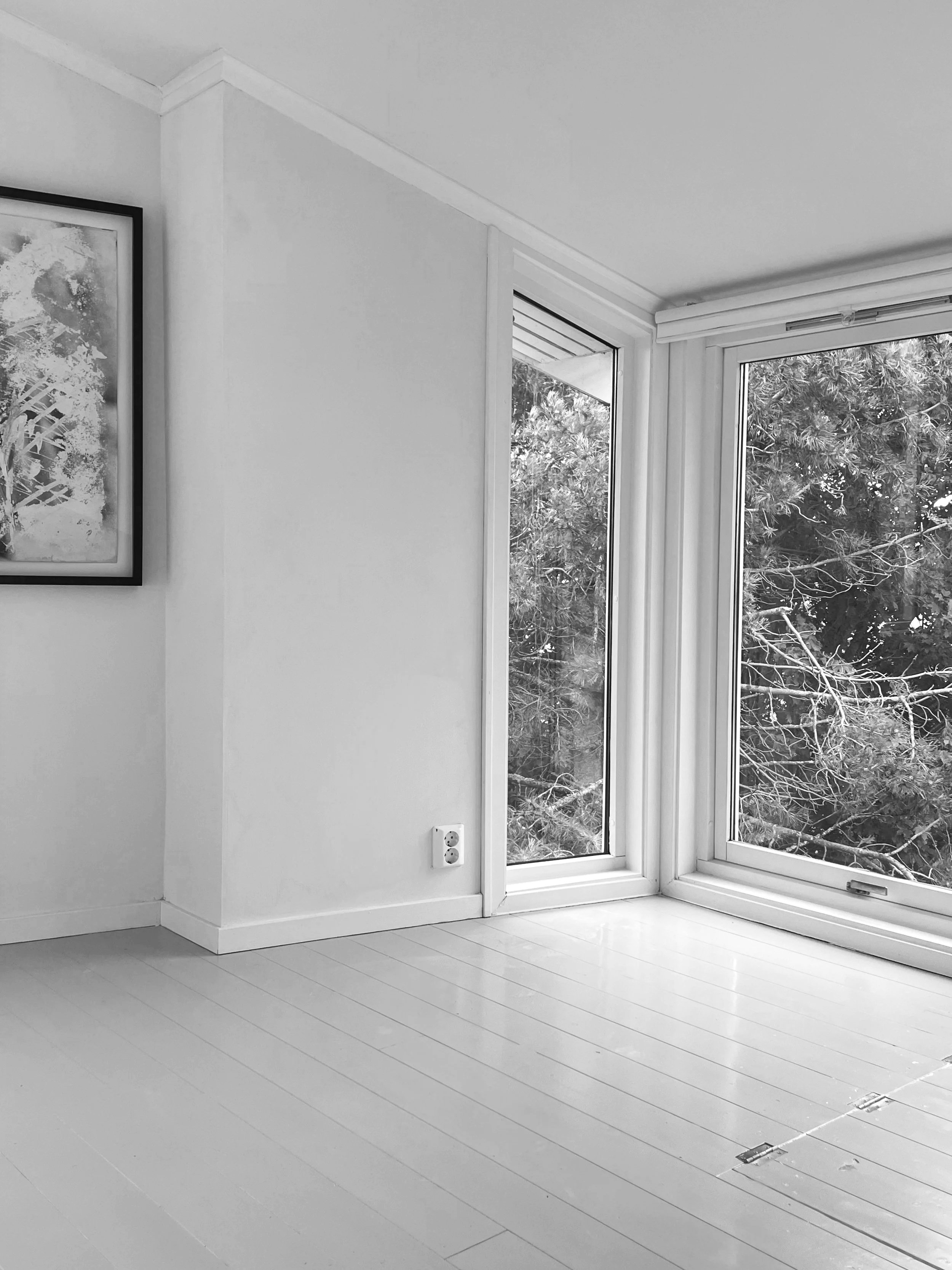 A corner of a room with white walls and light-colored wooden flooring, large windows showing trees outside, and a framed artwork on the wall.