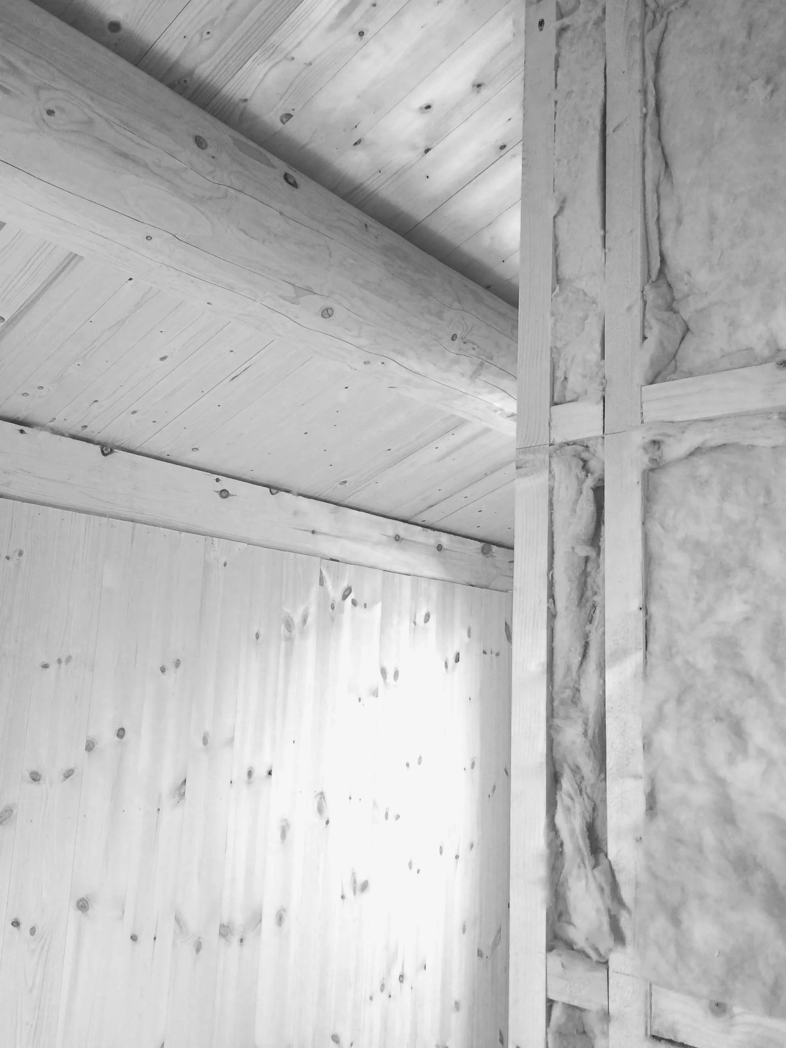 Interior view of a construction site showing wooden beams, plywood walls, and insulation in a black-and-white photo.