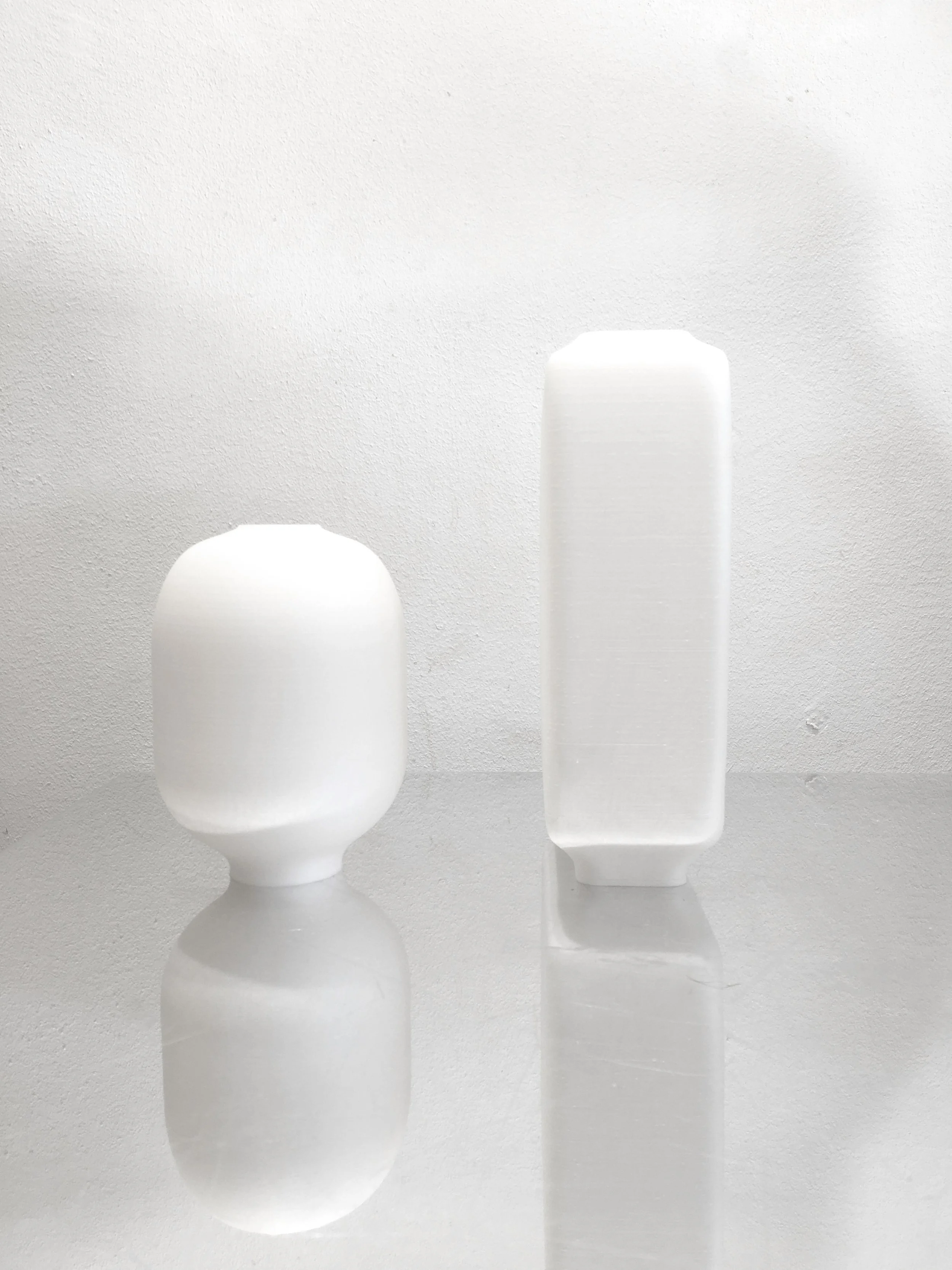 Two white modern vases with minimalist design on a reflective surface against a textured white wall.