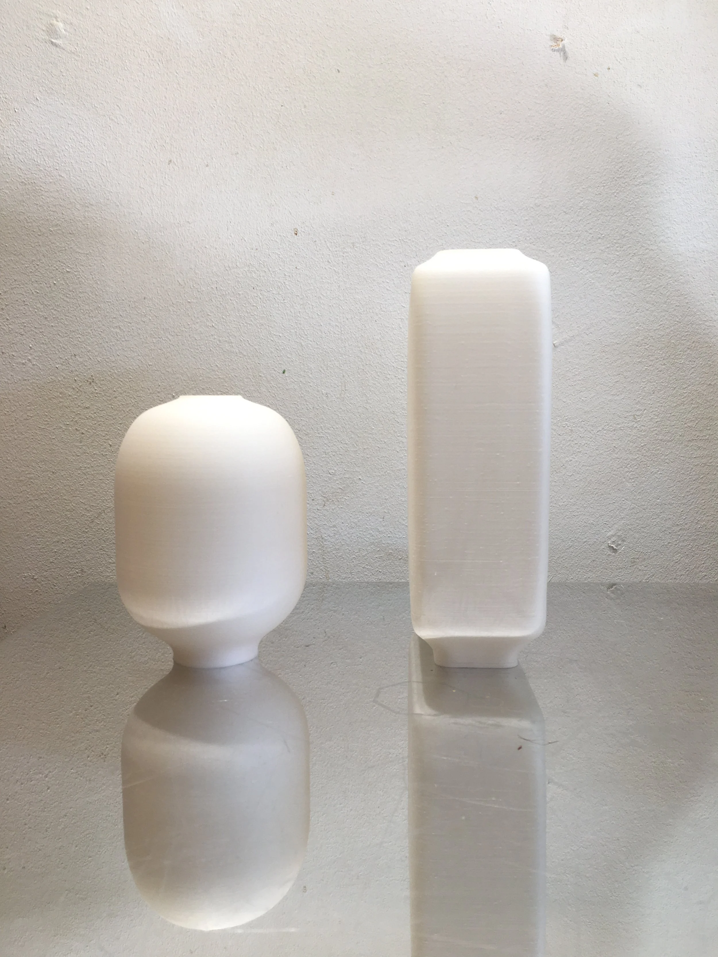 Two white abstract sculptural vessels on a reflective surface against a textured white wall, one rounded and one rectangular.