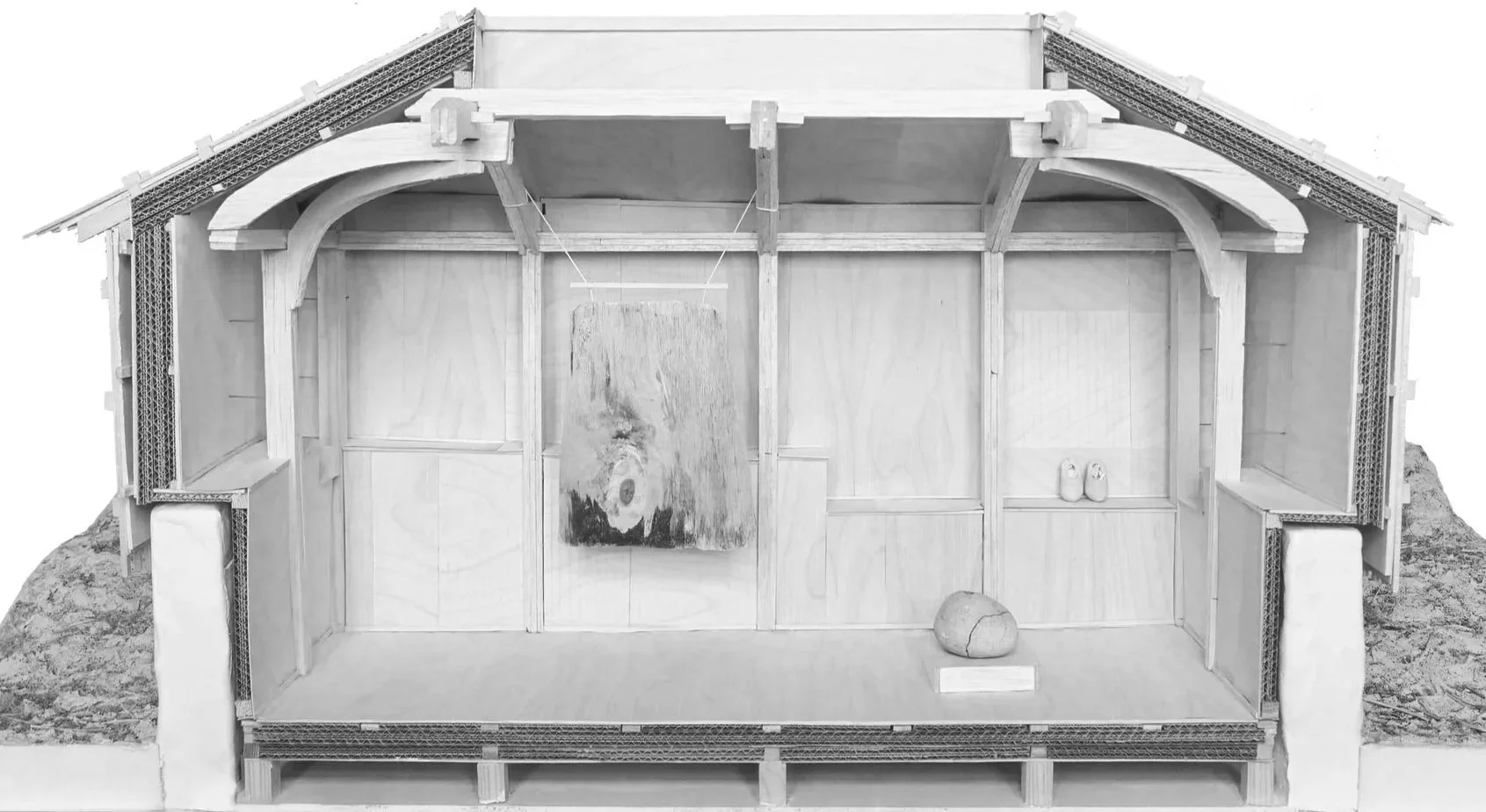 A cross-section of a model house made from cardboard, showing the interior walls, roof, and structural framework, with objects like a board, rocks, and shoes inside.