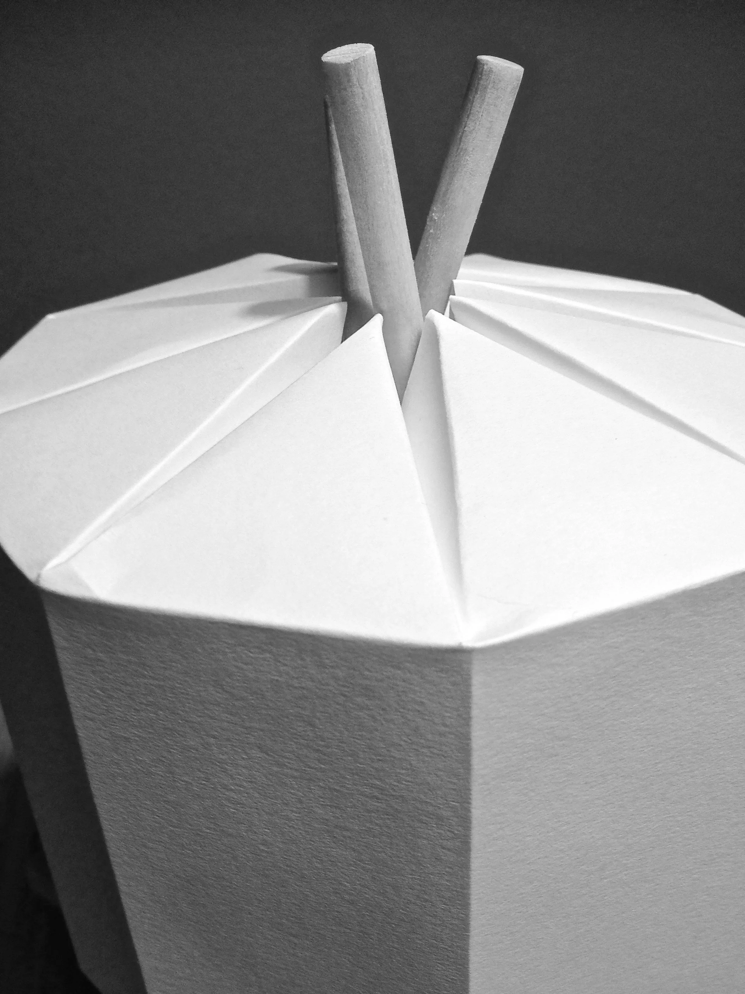 A close-up of a white paper box with dented sides and an open top, containing two wooden sticks sticking out from the center.