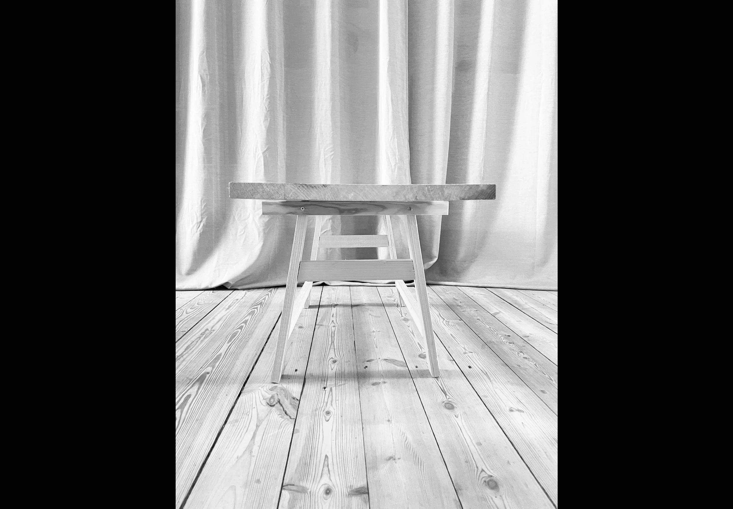 A black and white photo of a simple wooden table with a curtain backdrop and wooden floor.