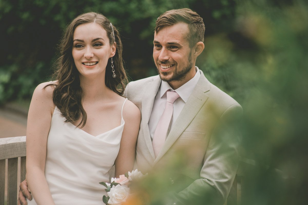 Smiling couple in formal attire, outdoors, with a woman in a white dress holding flowers and a man in a light suit with a pink tie.