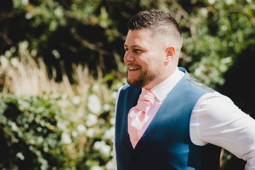 Man in blue suit vest and pink tie smiling outdoors with blurred foliage background.