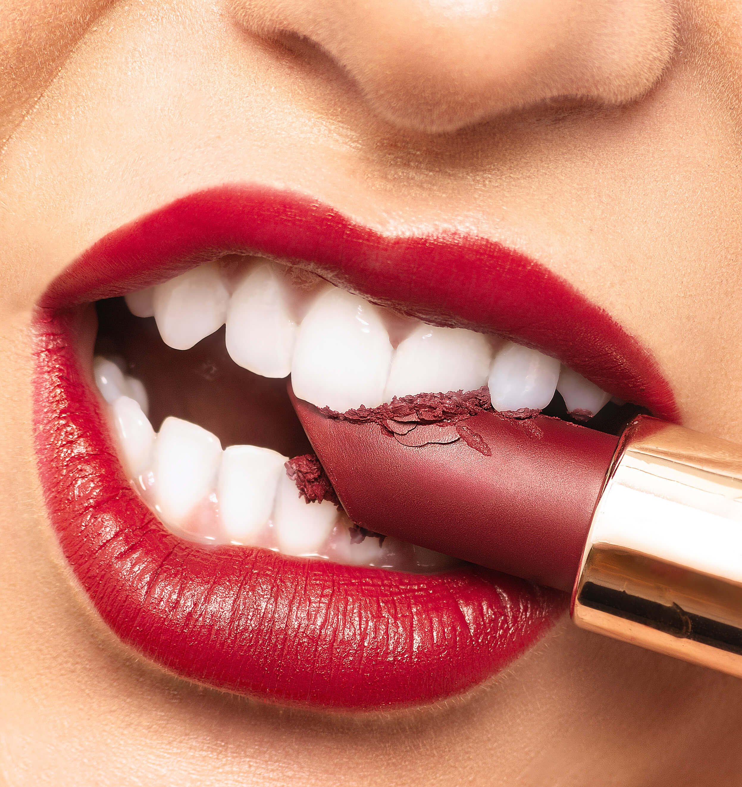 Close-up of a person biting a red lipstick with red lipstick on their lips, showing white teeth.