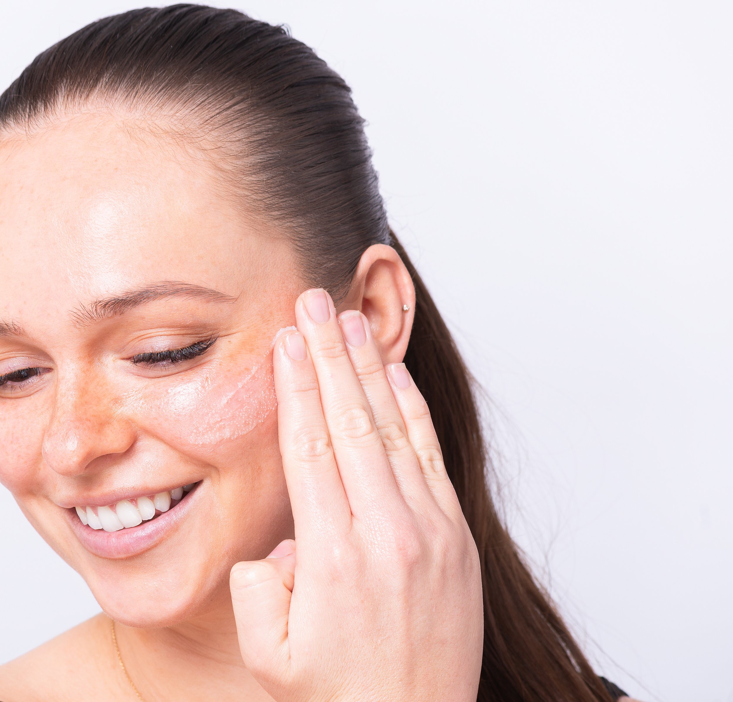 Woman applying facial cream to her cheek with a smile, demonstrating skincare routine.