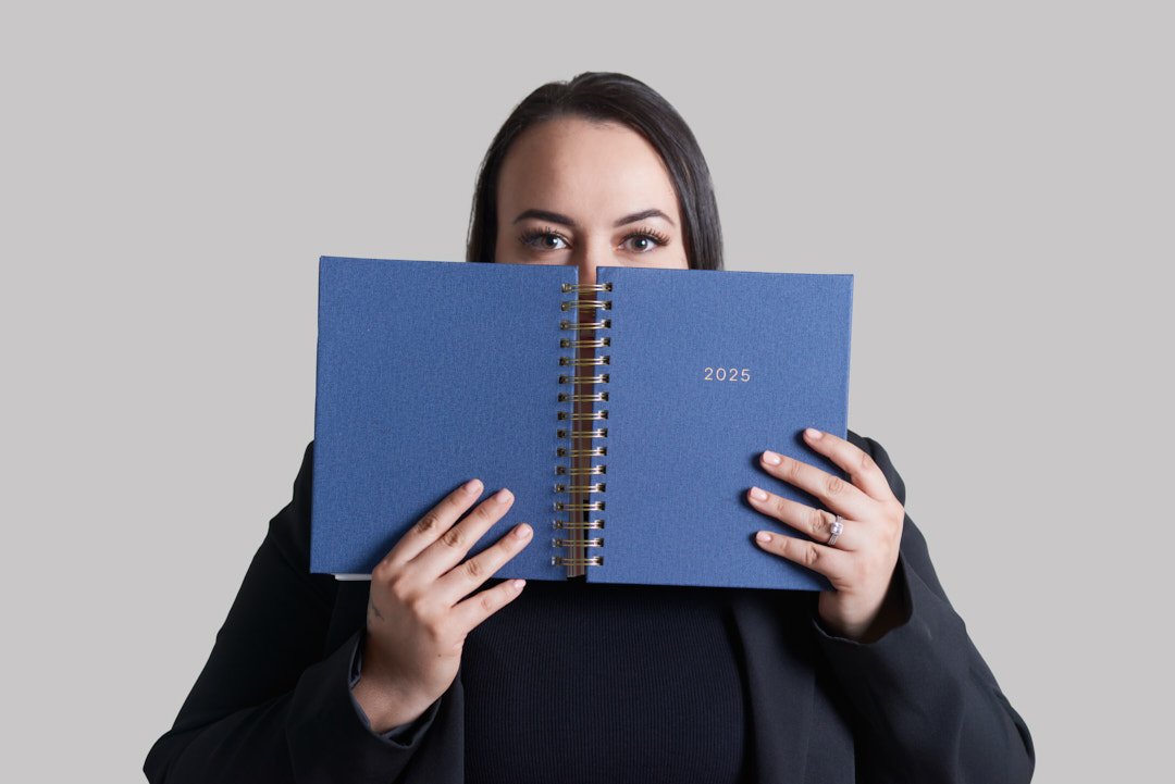 Woman holding a blue 2025 planner in front of her face, partially covering her face, with only her eyes and forehead visible.