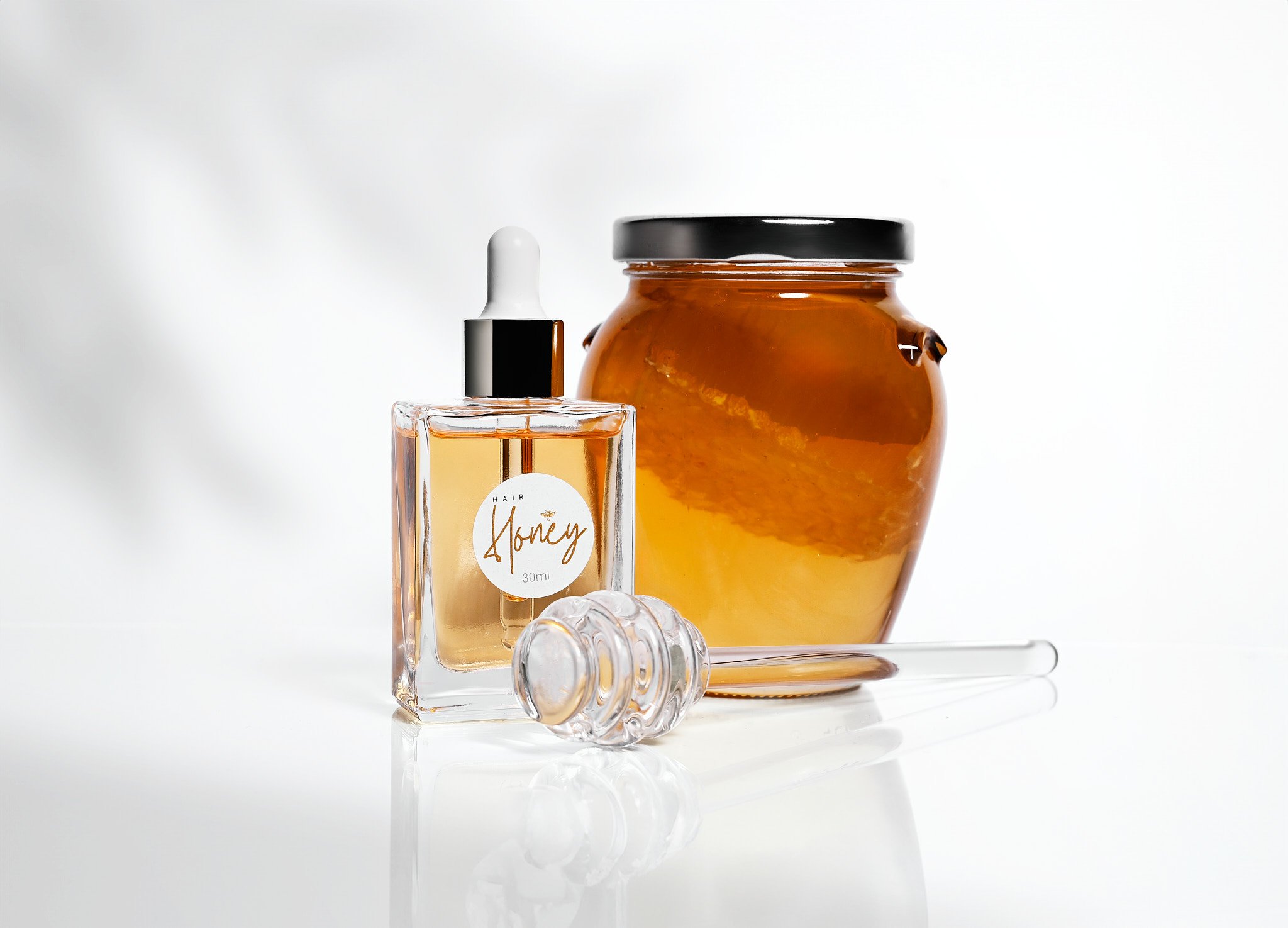 Glass jar of honey, clear dropper bottle labeled 'hair honey 30ml,' and a glass honey dipper on a white background.