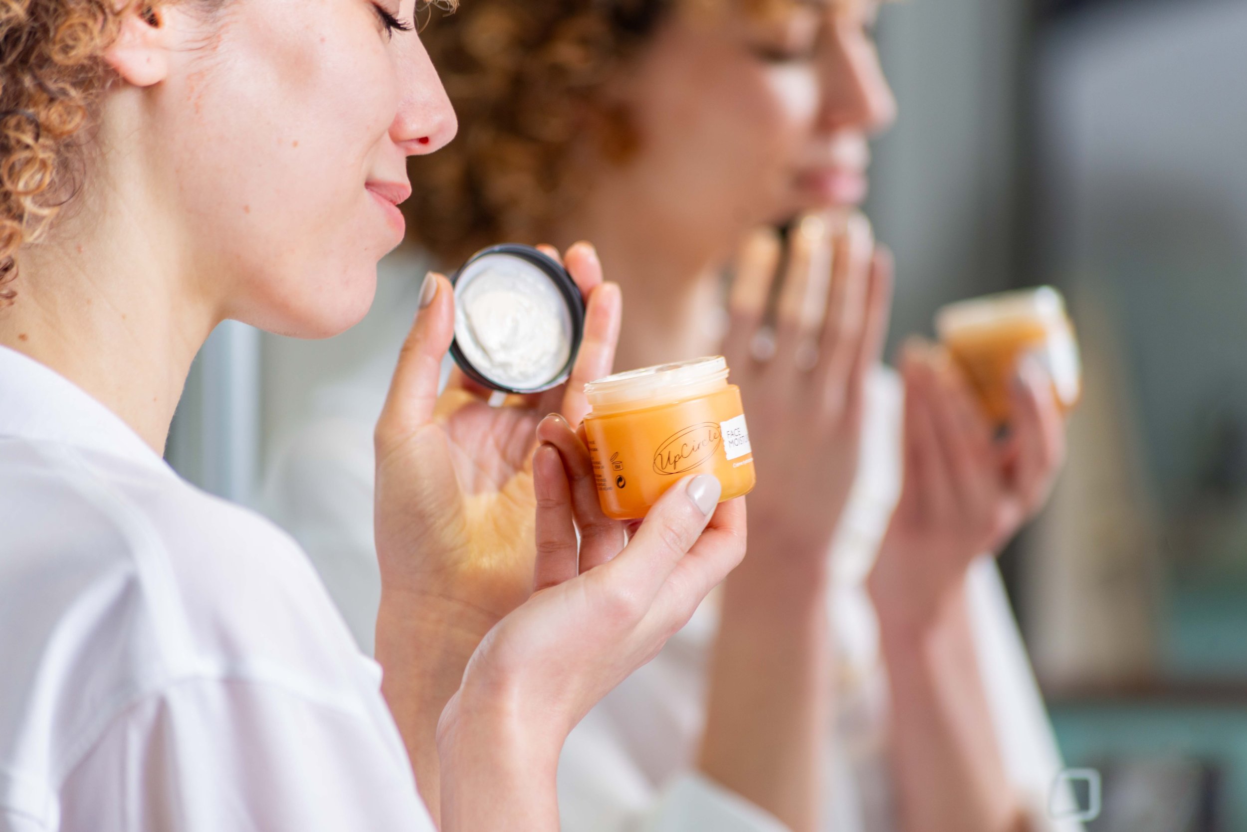 Woman applying face moisturizer in front of a mirror.