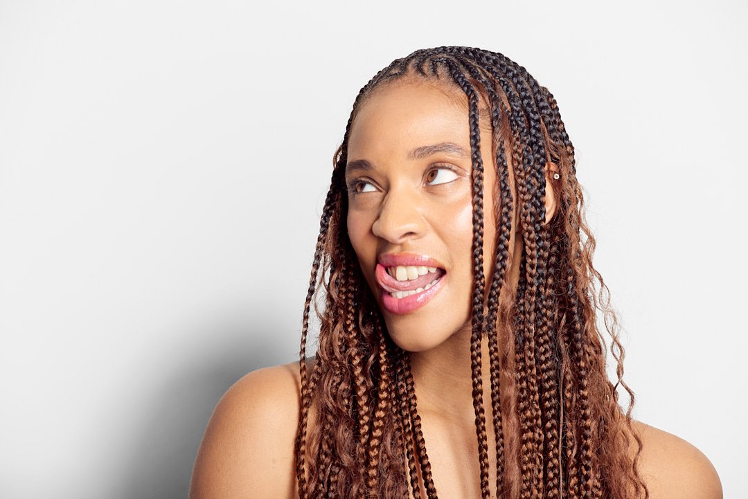 Person with braided hair looking to the side, sticking out tongue, against a plain background.