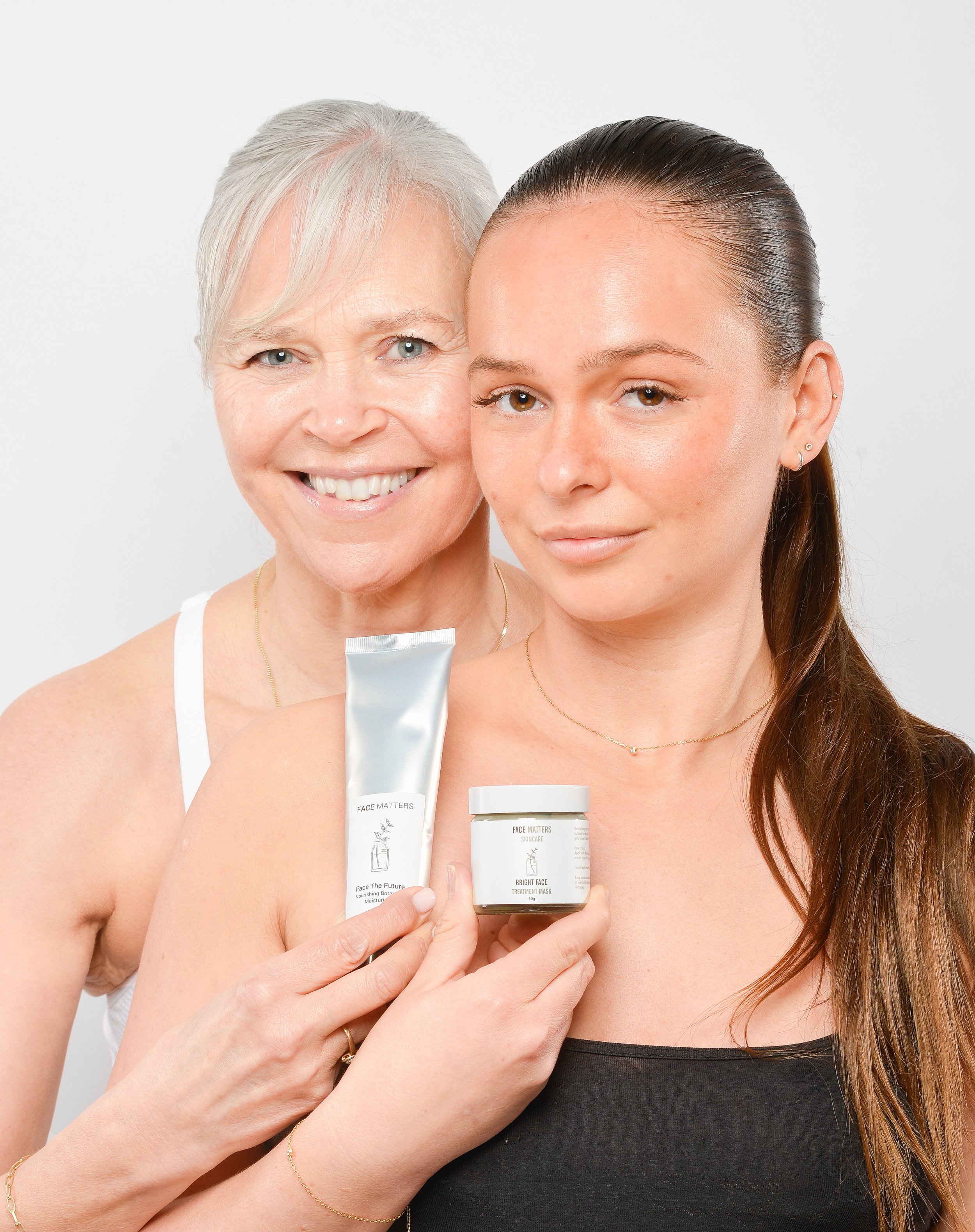 Two women holding skincare products, smiling, white background.