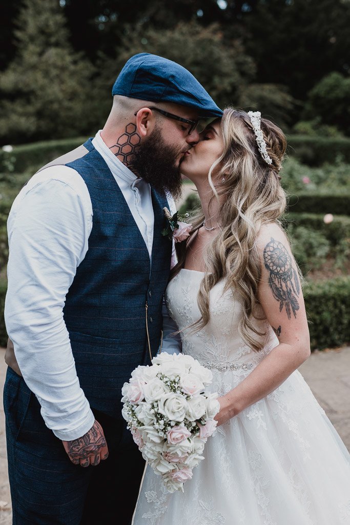 Bride and groom kissing outdoors, bride holding a bouquet of white and pink roses, groom wearing a blue suit and cap.