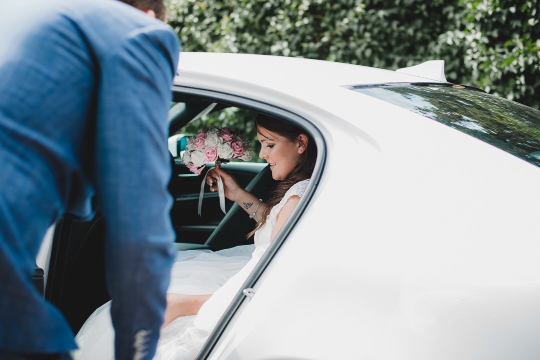 A bride in a white dress holding a bouquet of pink and white flowers, sitting inside a white car, with a person in a blue suit assisting her.