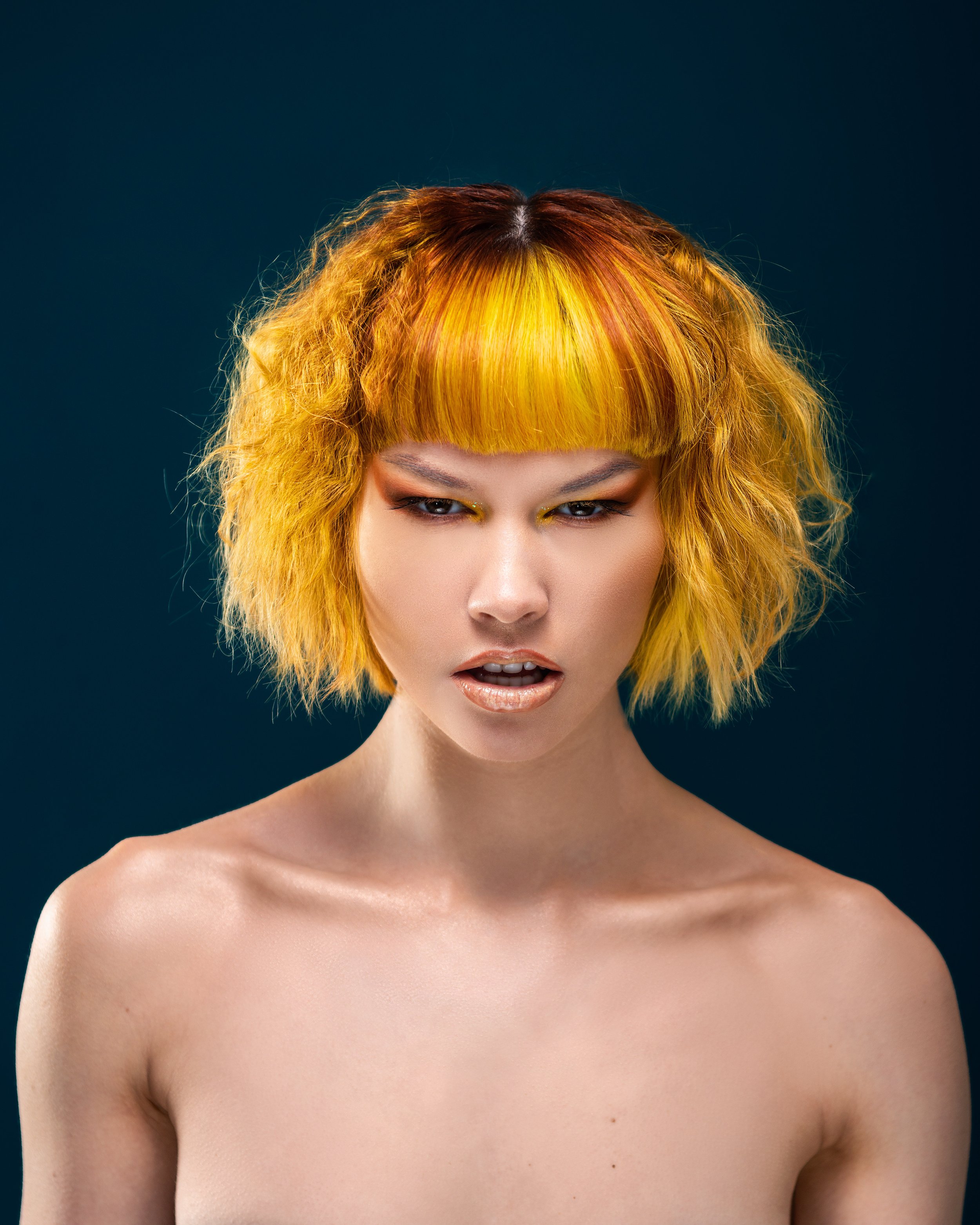 Person with orange and yellow wavy bob hairstyle and dramatic makeup on a dark background.