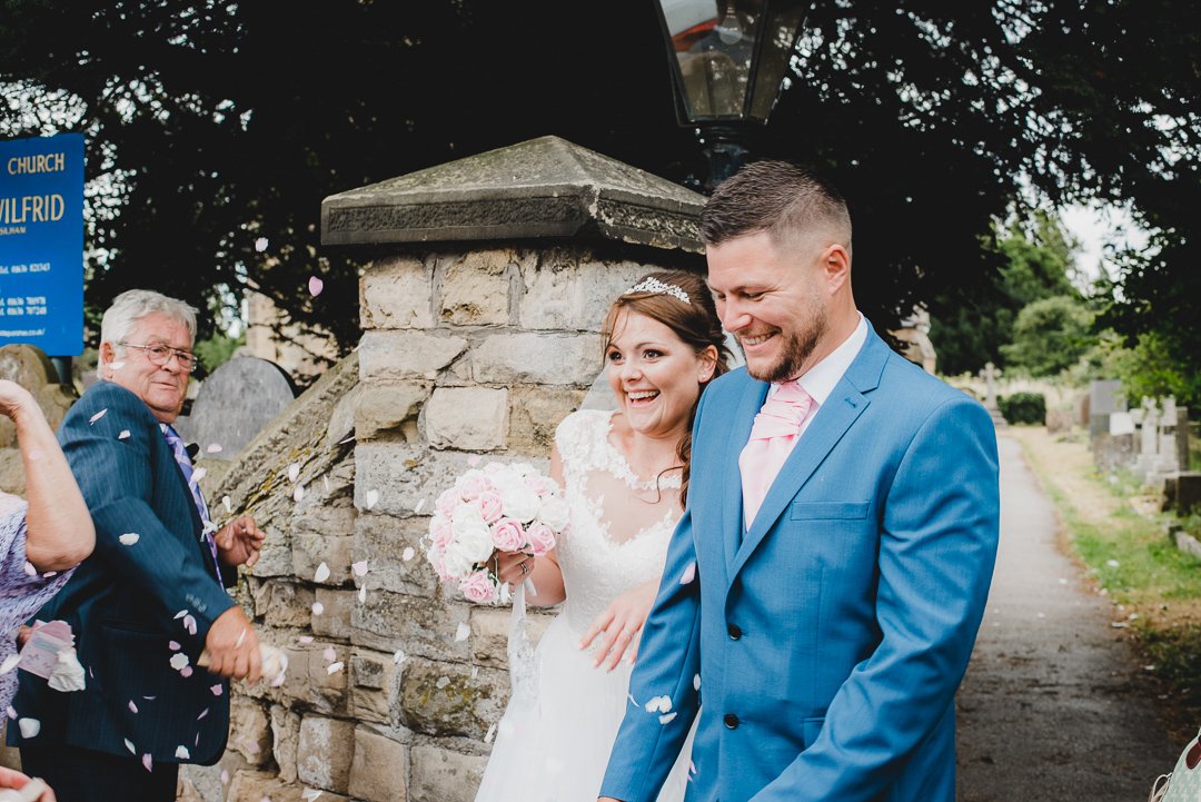 Bride and groom smiling with flower petals being thrown at them outside a church.