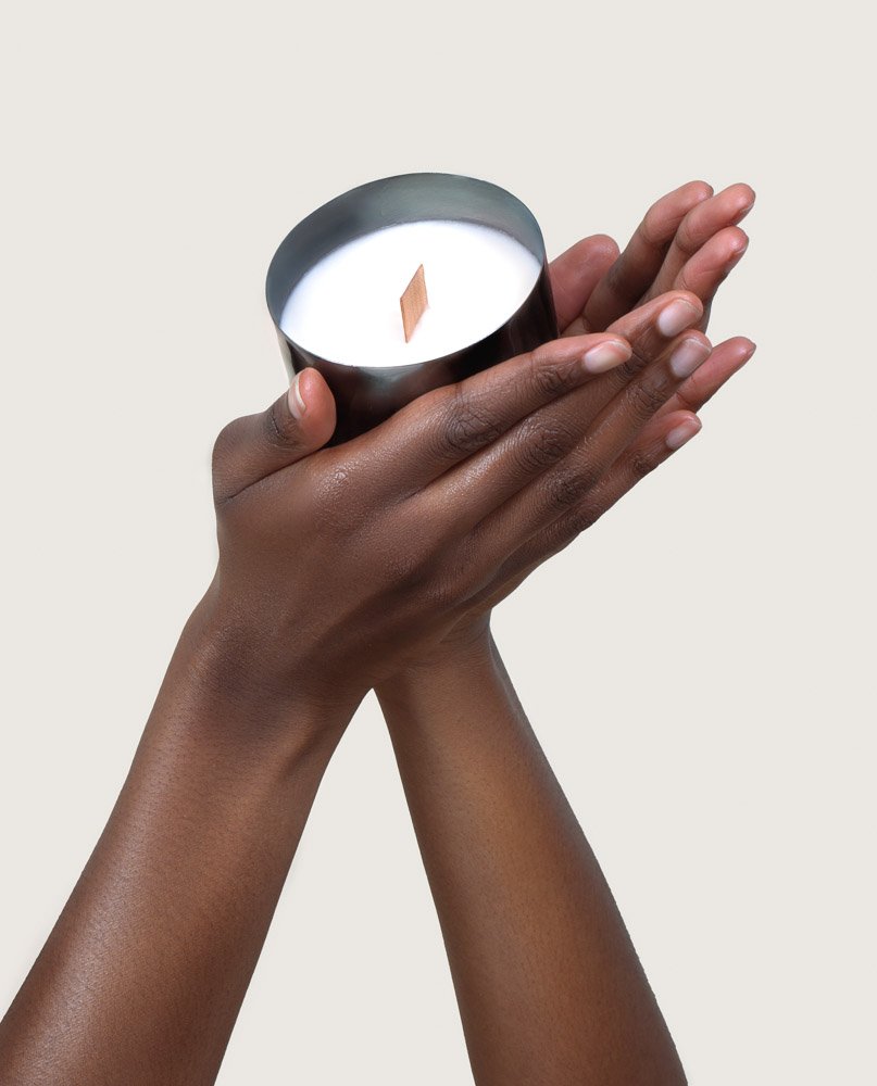 Hands holding a candle in a metal container with a wooden wick on a light background.