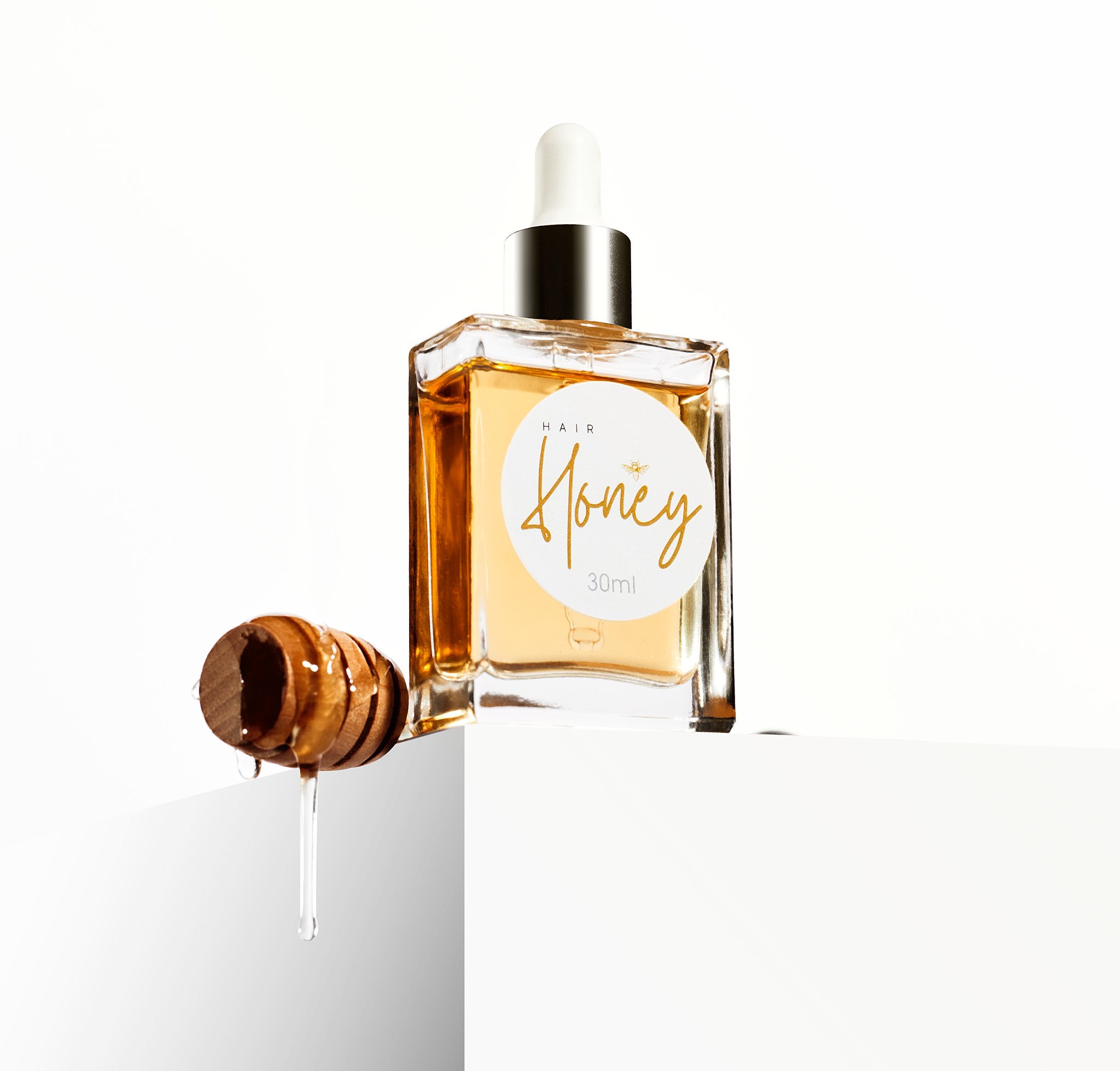 A bottle of hair oil labeled 'Hair Honey' with a wooden honey dipper dripping honey beside it.