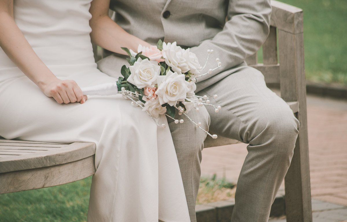 Bride and groom seated on a bench, holding a bouquet of white and pink flowers.